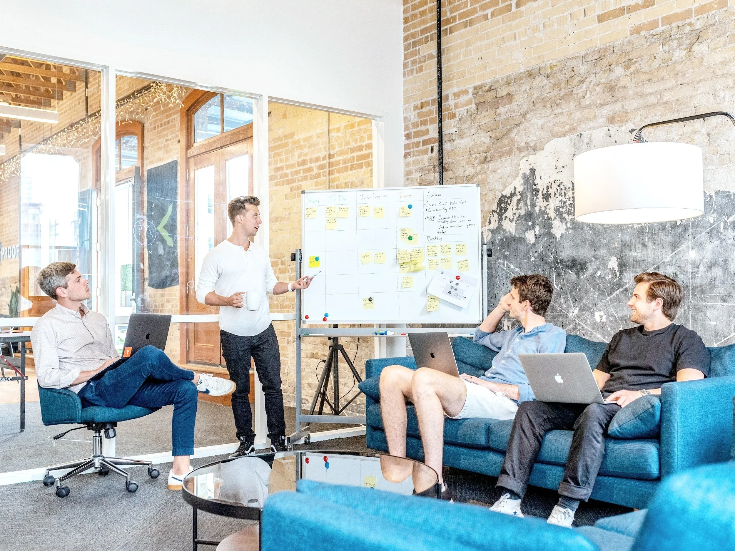 Four young men in a modern office space, one standing and presenting with a whiteboard, three sitting with laptops and listening.