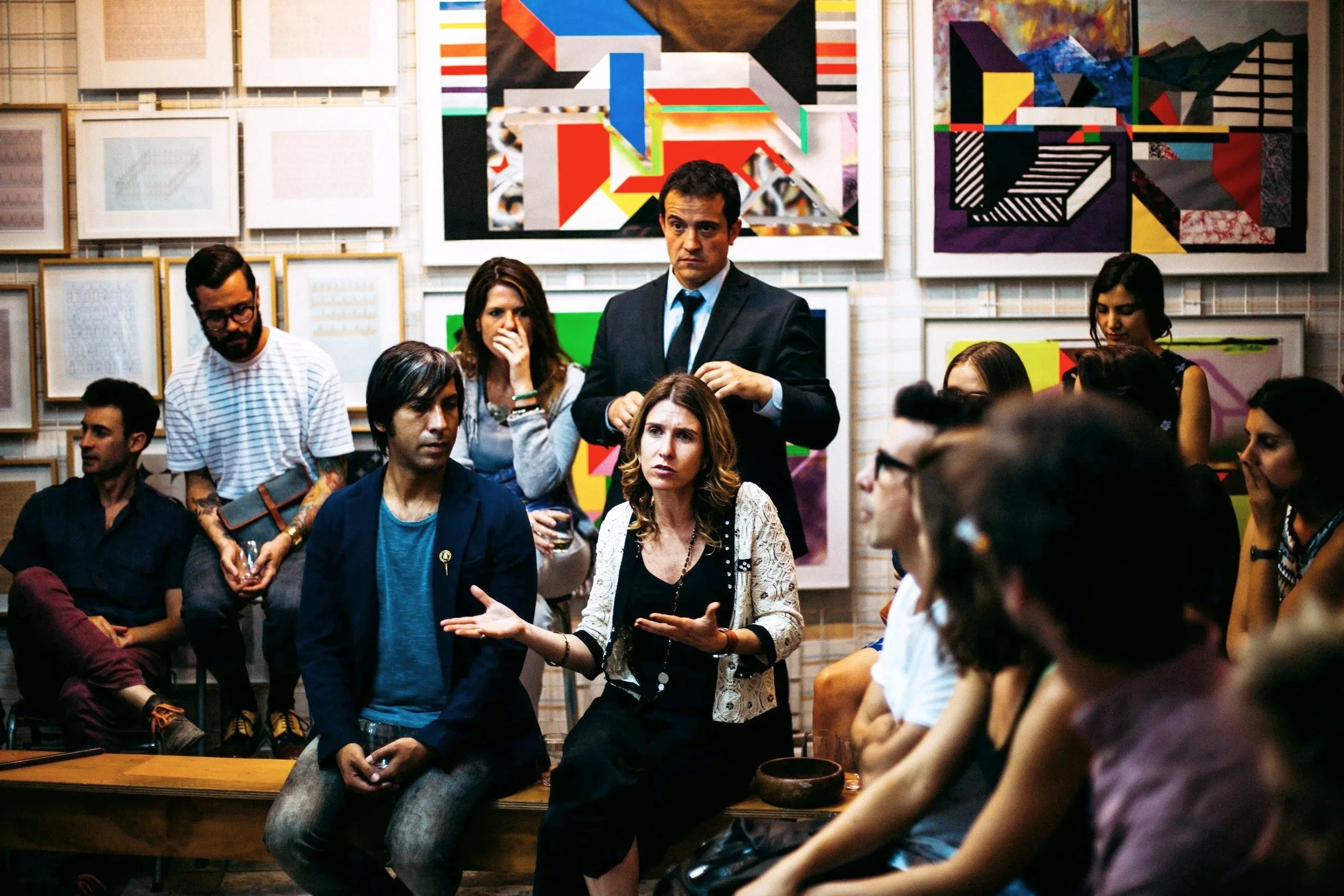 Group of people participating in a discussion or workshop in an art gallery or studio with colorful abstract paintings on the wall behind them.