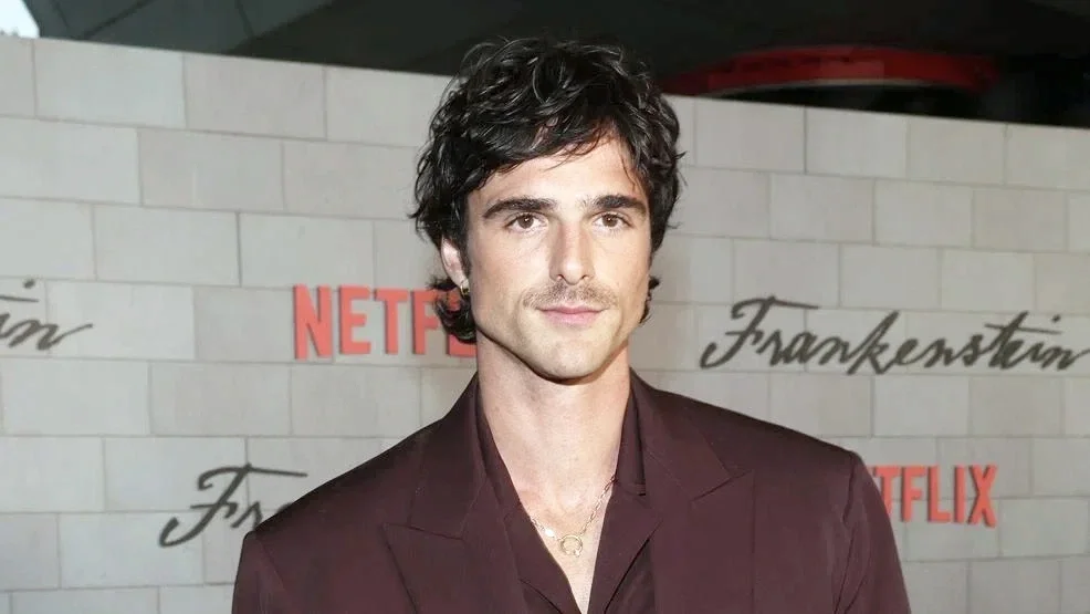 A man with dark wavy hair and fair skin posing in front of a backdrop with the Netflix logo and the word 'Frankenstein' written in cursive.