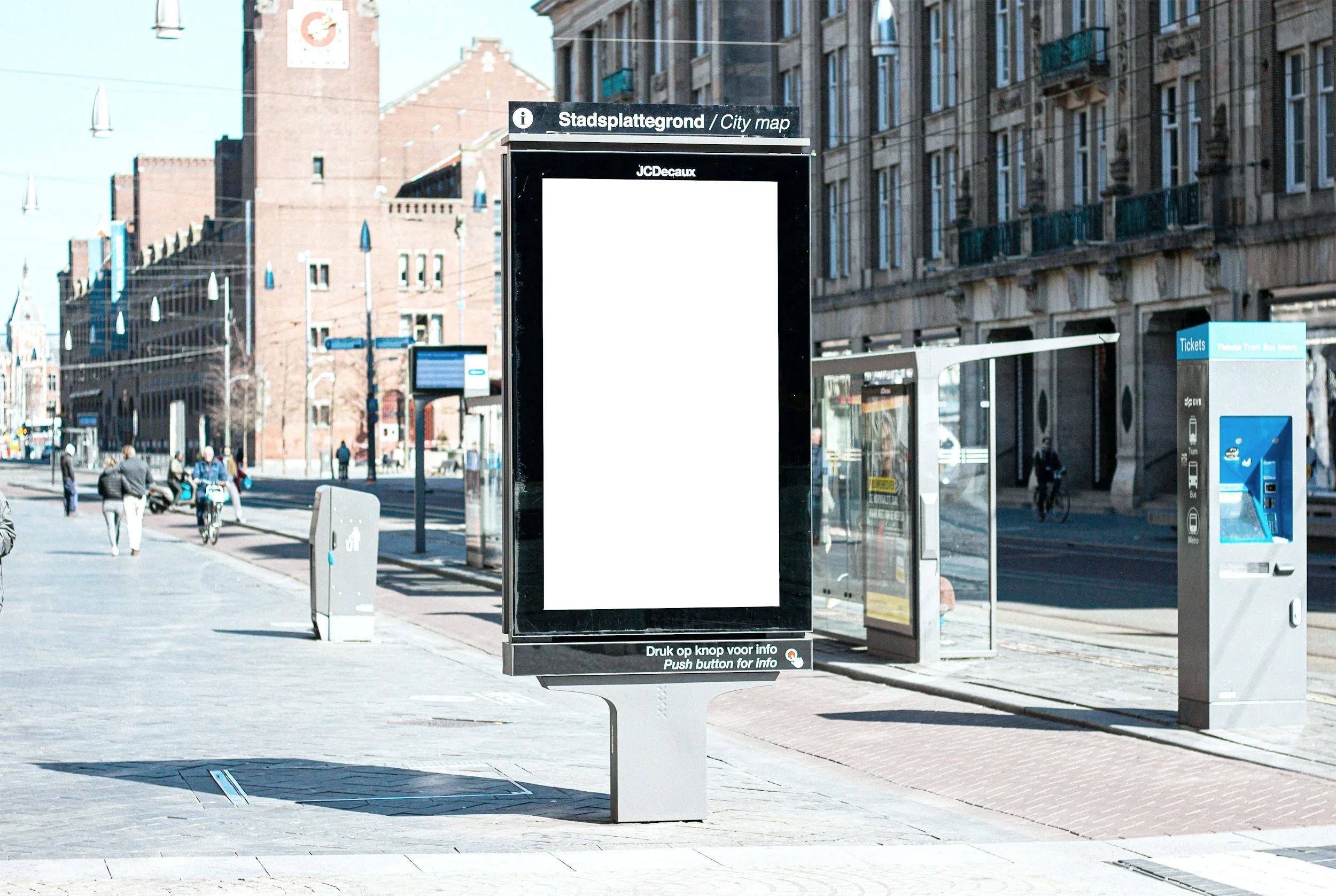 City street scene with digital information kiosk in the foreground, a bus ticket machine on the right, sidewalk, pedestrians, buildings, and blue sky.