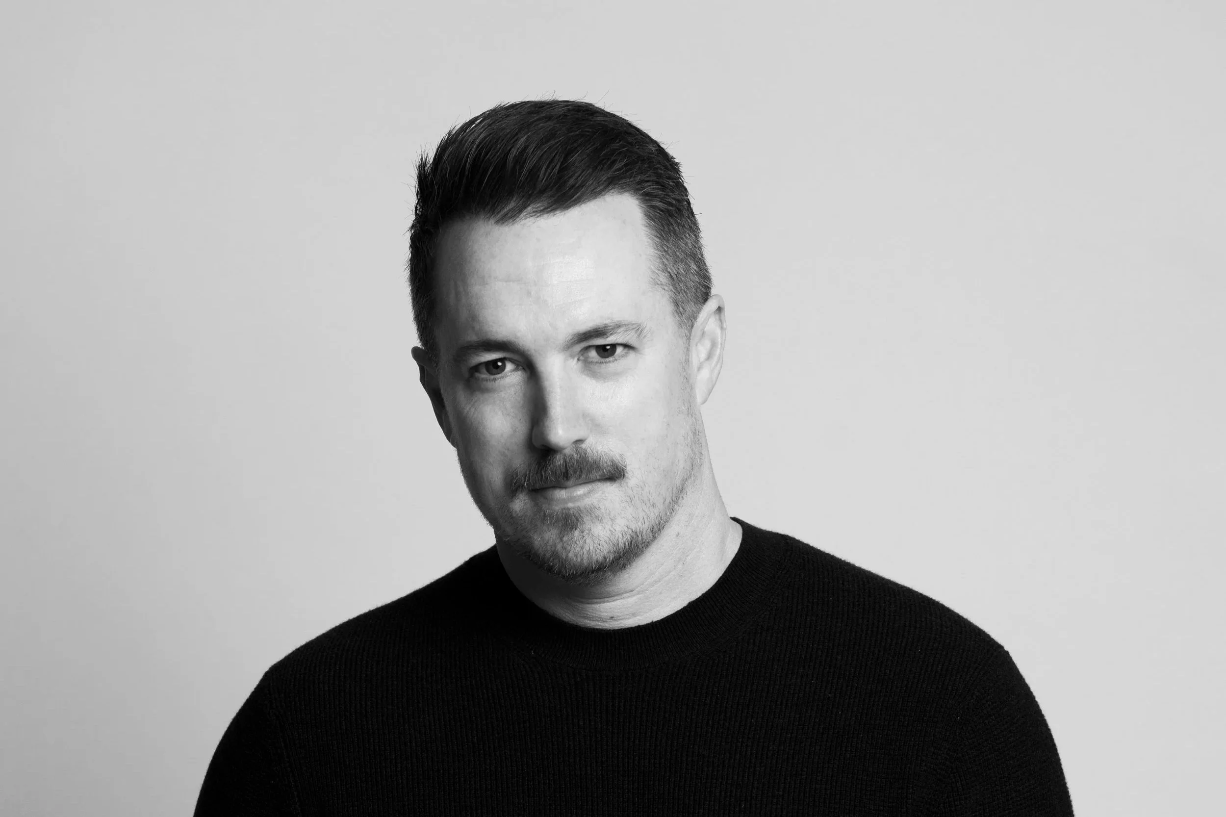 Black and white portrait of a man with short dark hair, mustache, and beard, wearing a black sweater, looking at the camera against plain light background.