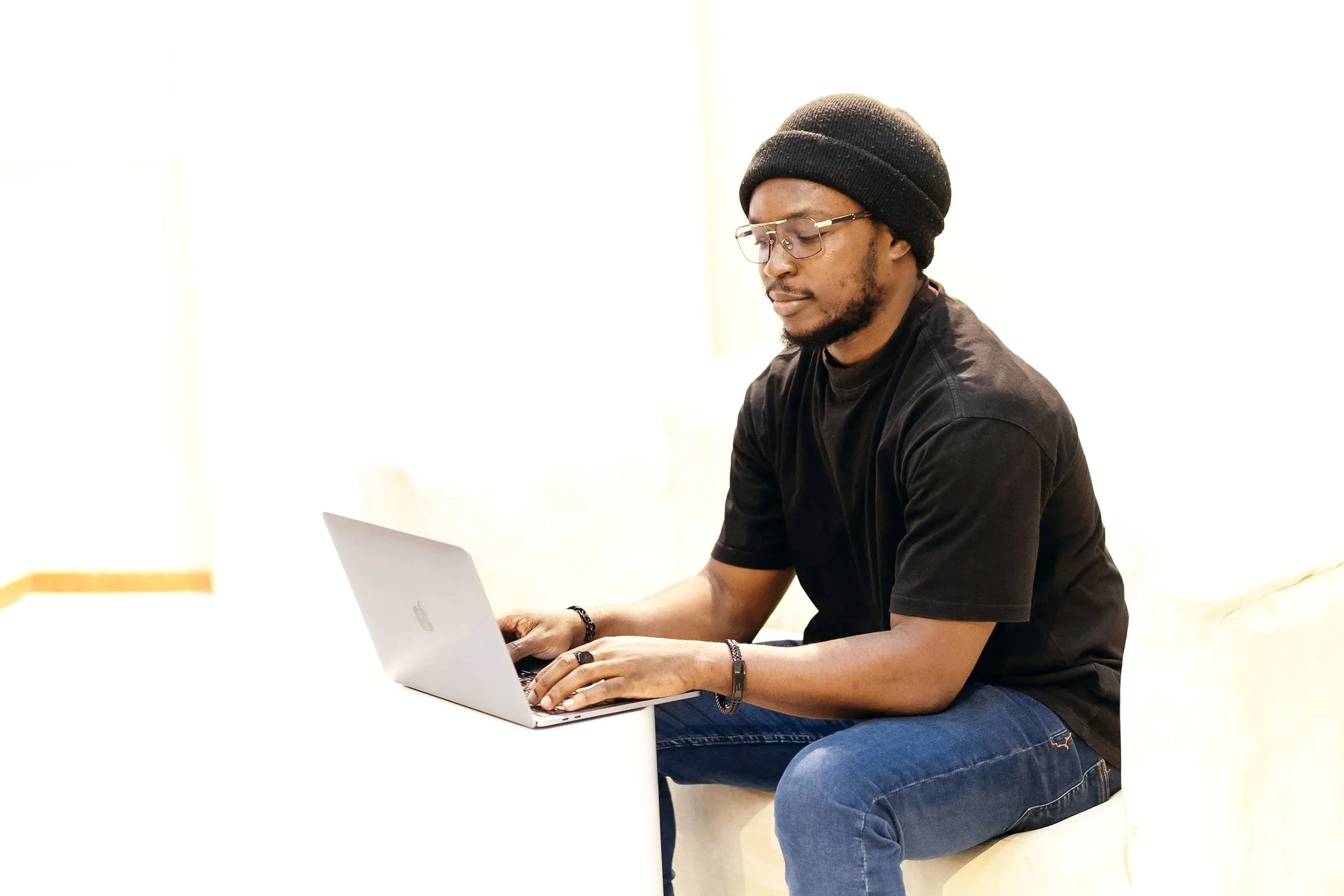 A young man wearing glasses, a black beanie, and black t-shirt working on a silver laptop while sitting on a white couch in a bright room.