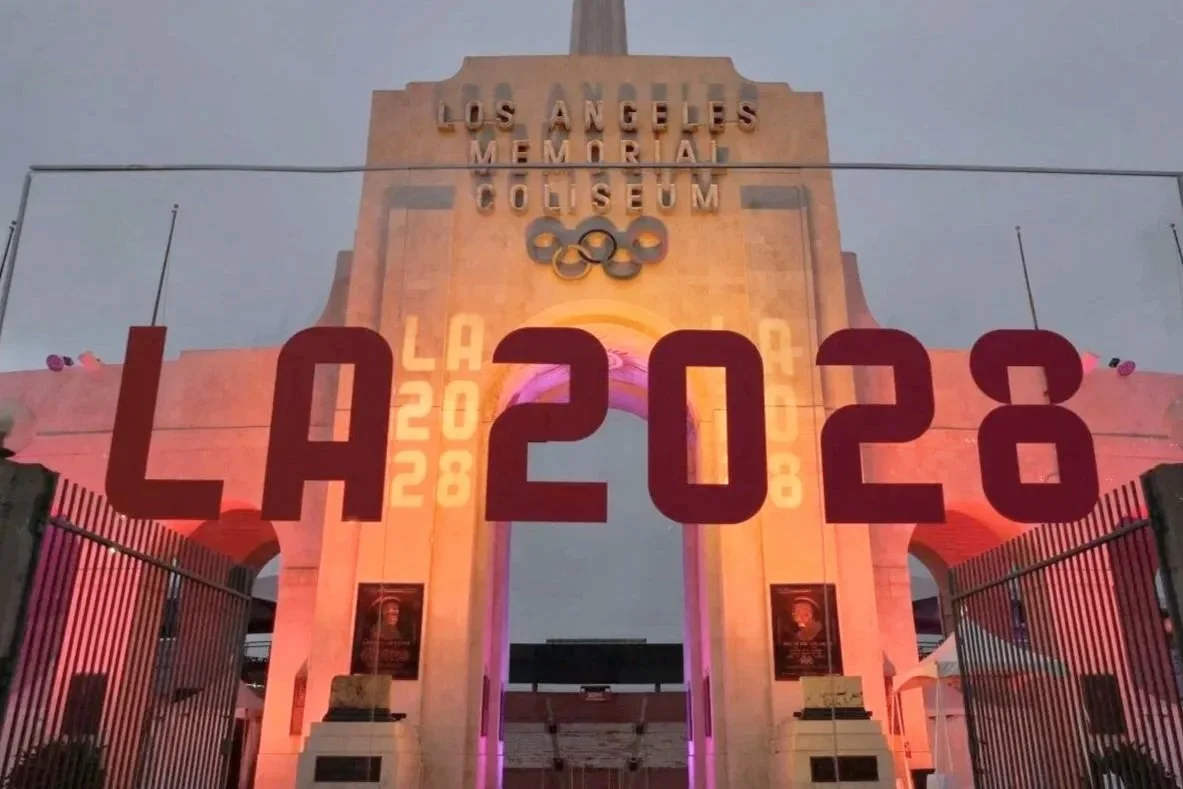 Entrance arch to the Los Angeles Memorial Coliseum illuminated with pink and purple lights, with the date 'LA 2028' and the year 2023 prominently displayed.