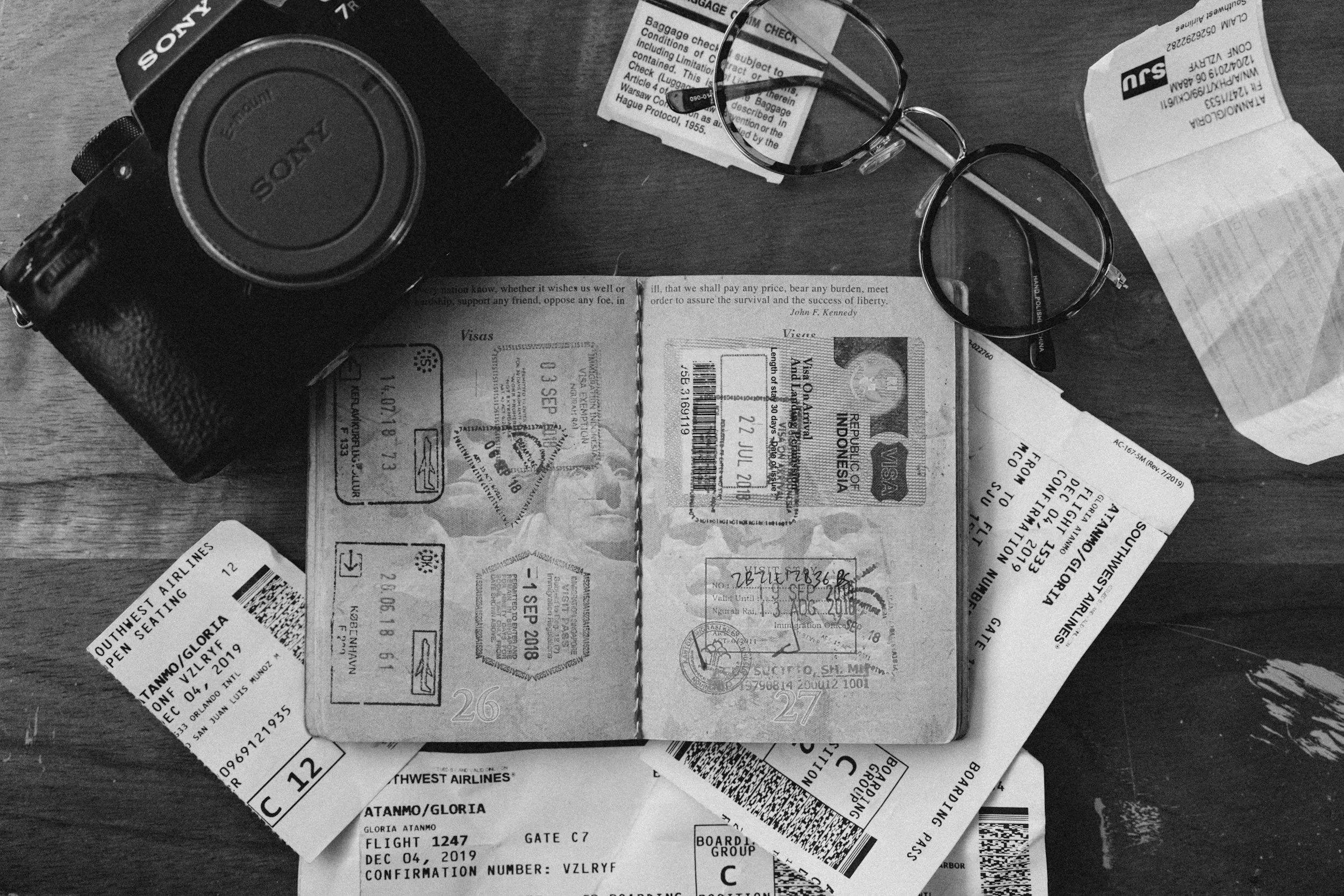 Black and white photograph of a travel scene with a camera, eyeglasses, various airline tickets, and a passport laid out on a wooden surface.