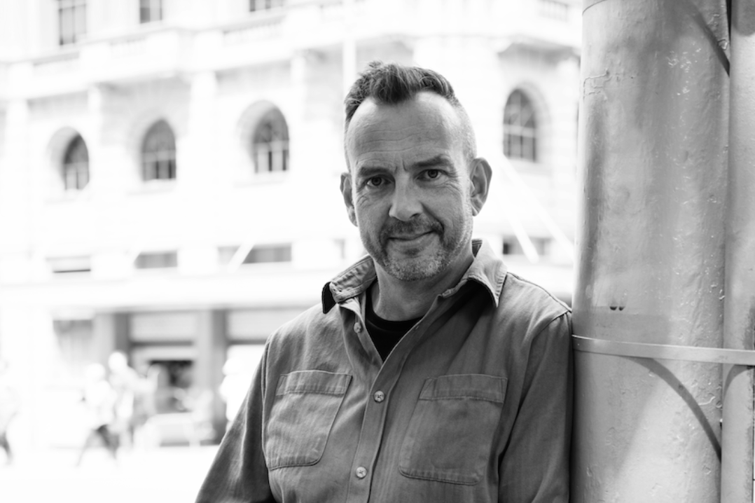 A man with short hair and a beard, wearing a casual button-up shirt, leaning against a pole on a city street.