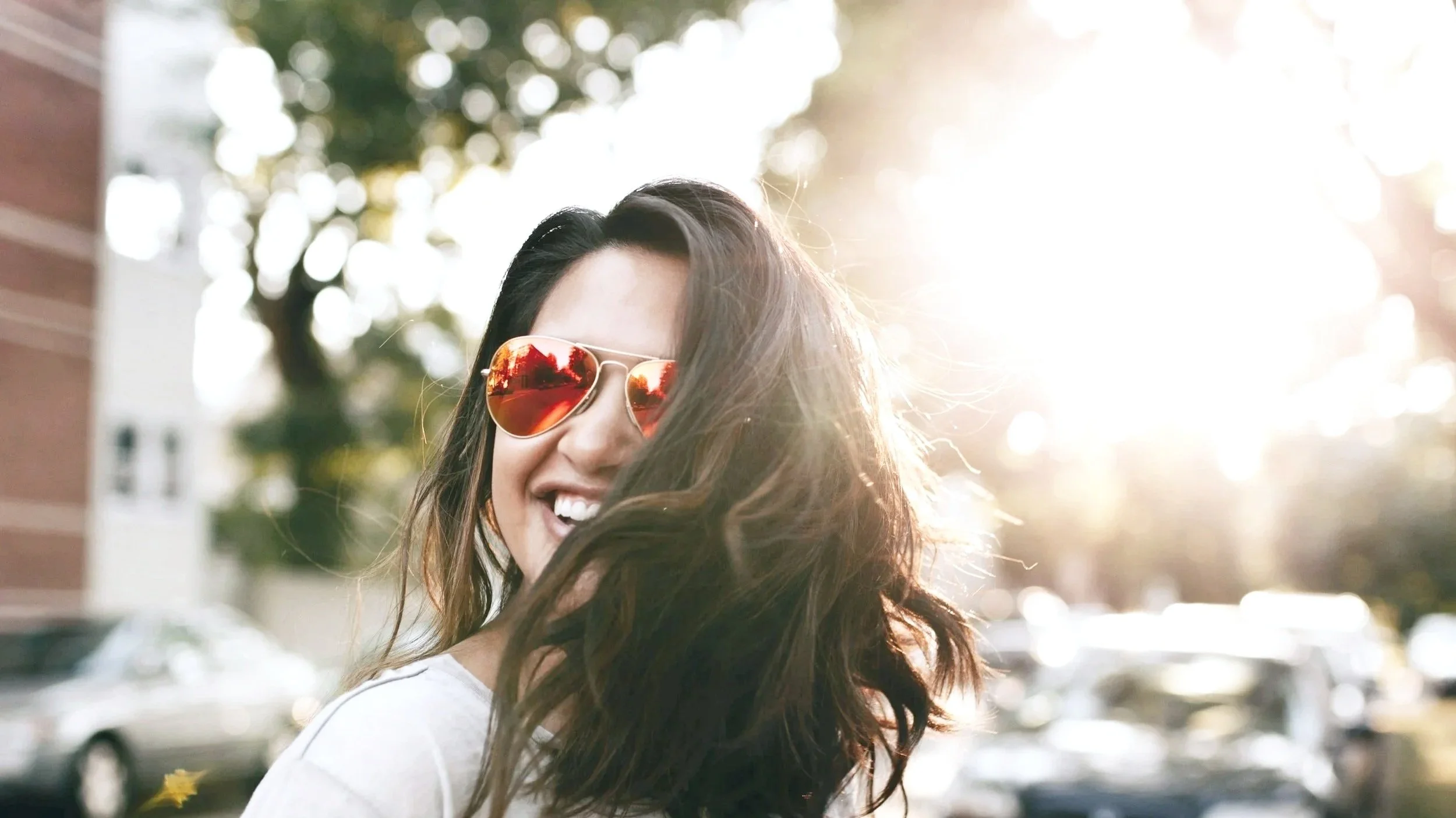A woman with long hair wearing red-tinted sunglasses, smiling and turning her head in front of a sunny, outdoor background with trees and parked cars.