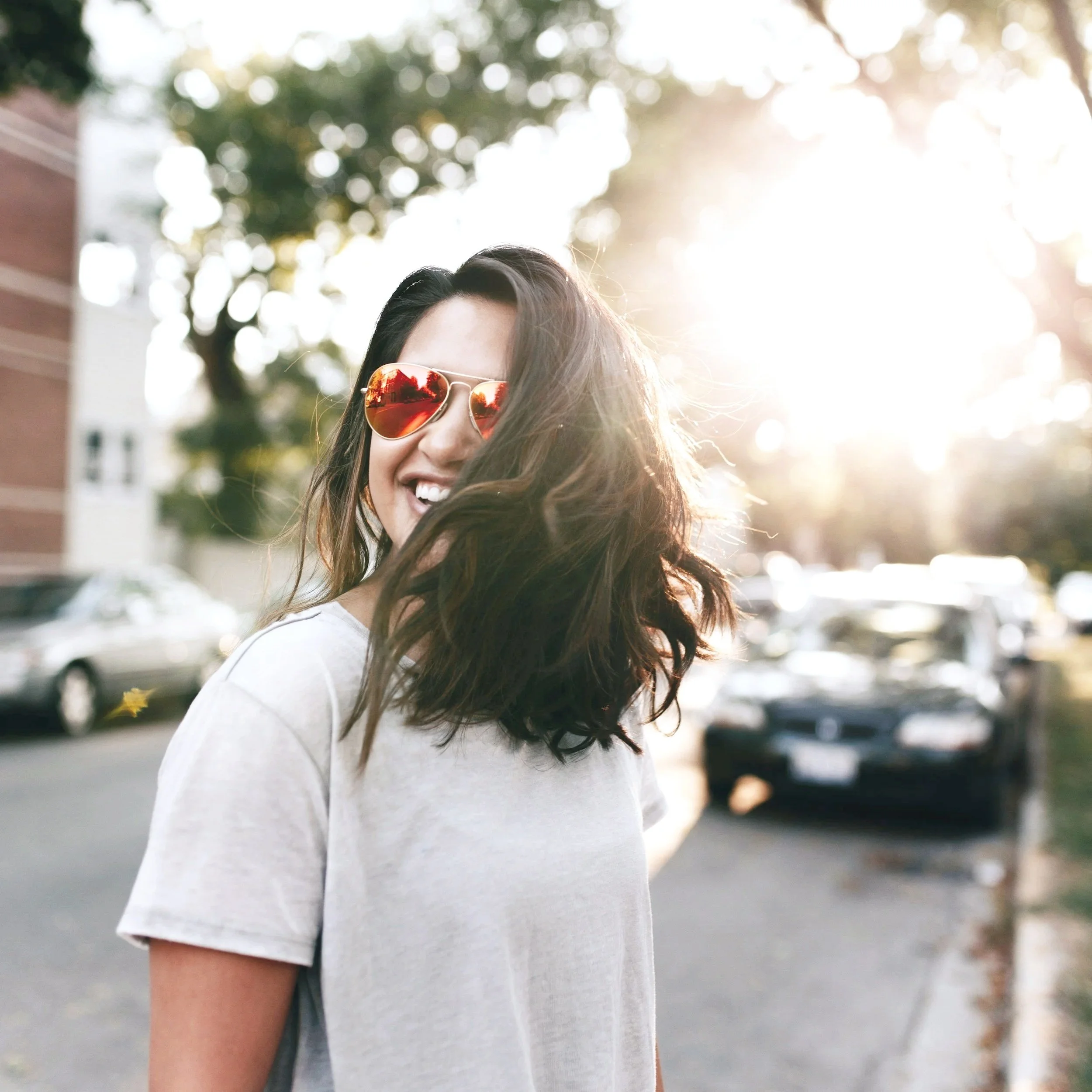 A woman with long dark hair wearing sunglasses smiling outdoors with cars and trees in the background, sunlight shining behind her.
