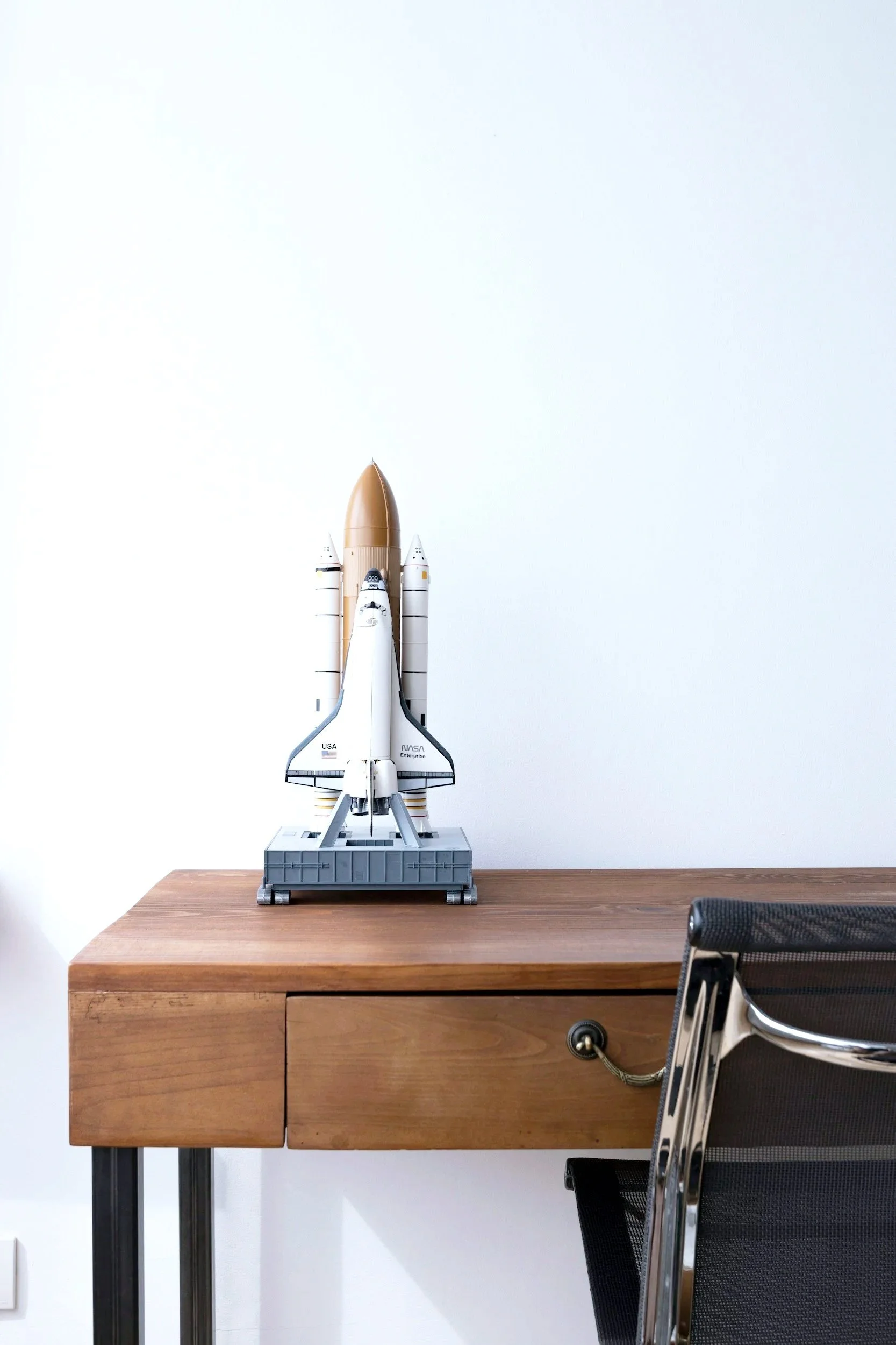 Model of a NASA space shuttle on a wooden desk with a plain white wall background.