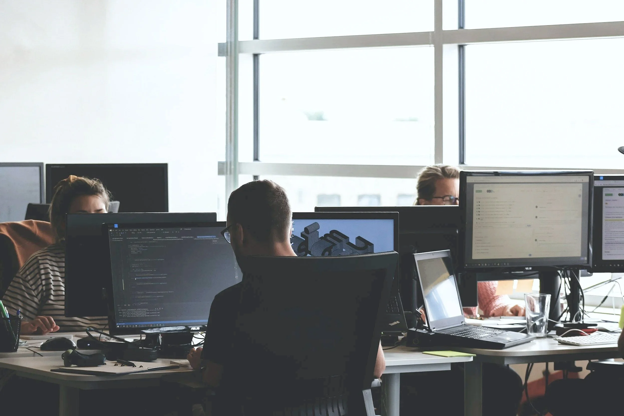 People working on computers in an office with large windows.