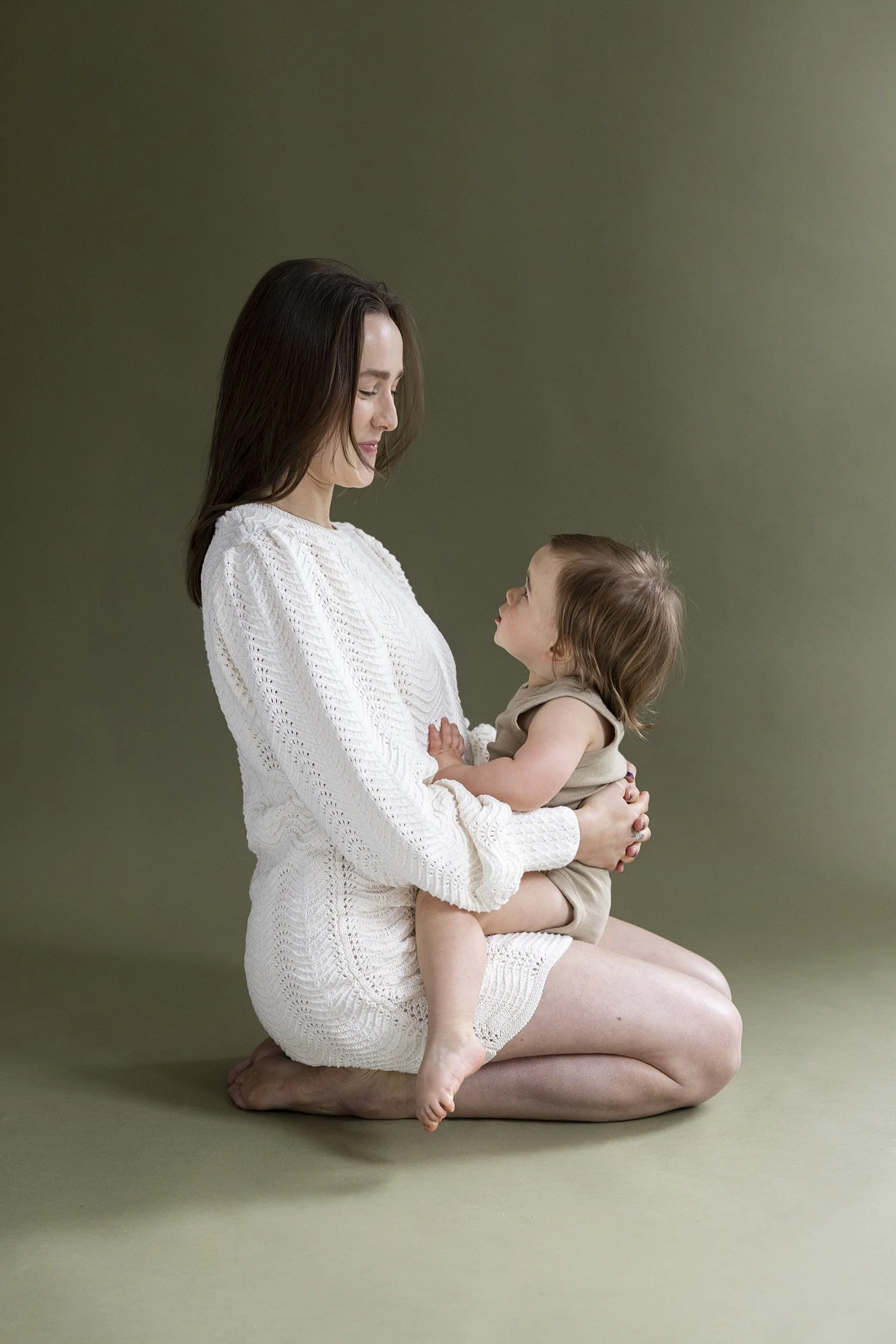 Baby sleep consultant in NYC sitting on the ground with her child in her lap in front of a green backdrop