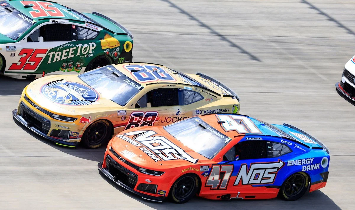 Three stock cars racing on a track, numbered 35, 88, and 47, with sponsor logos and colorful designs.
