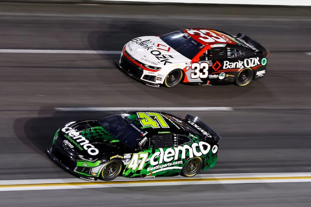 Two race cars driving on a racetrack at night. The car in front is black and green with the number 47, sponsored by Clemco. The car behind is black, white, and red with the number 33, sponsored by Bank OZK.