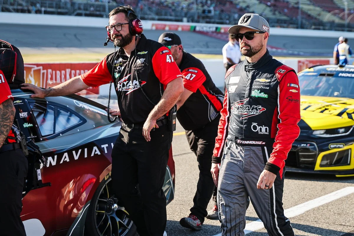 Two men in racing suits and one man in team uniform standing next to race cars at a racetrack.