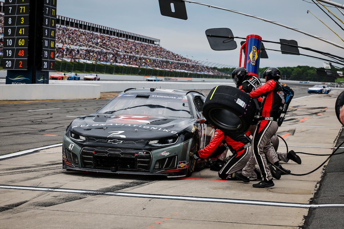 Race car pit stop with crew members changing tires on a black Chevrolet racing car at a motorsport event, with a crowded grandstand in the background.