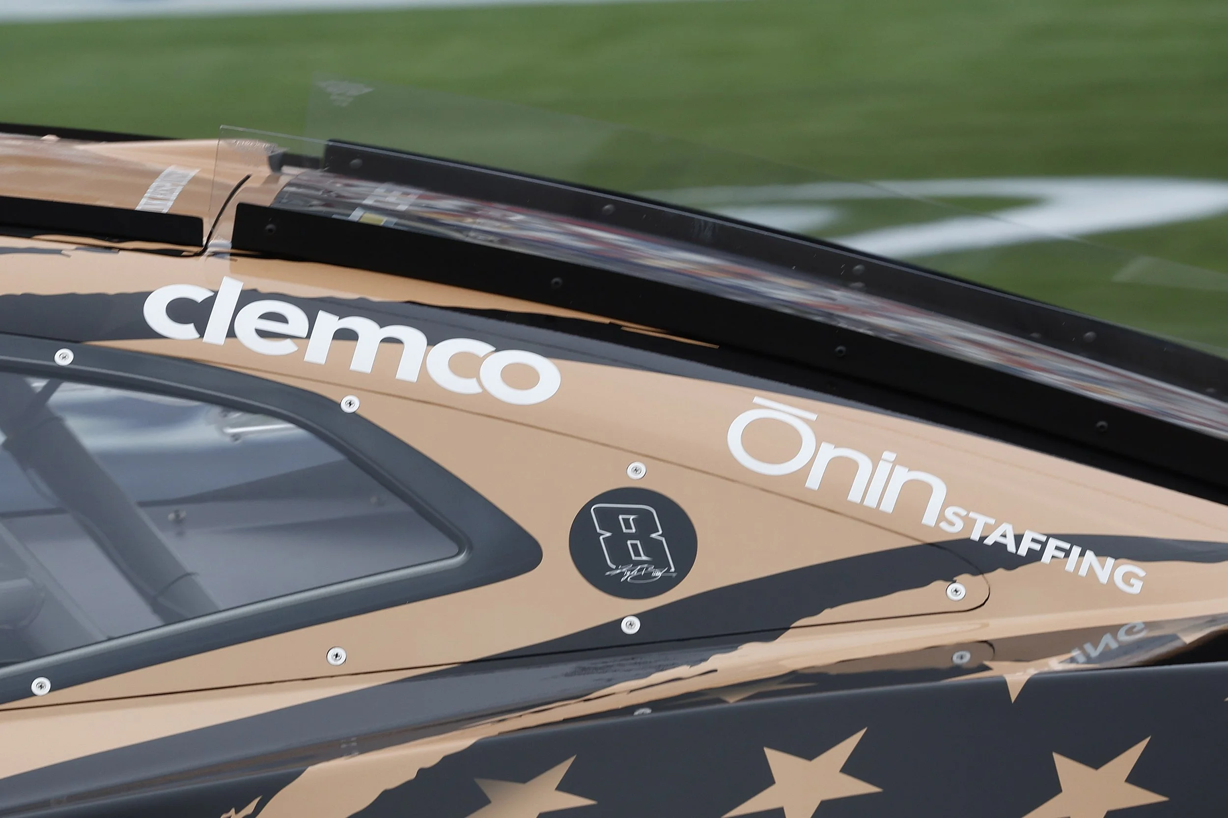 Close-up of a racing vehicle with sponsor decals, featuring the word 'clemco' in white letters, on a green racing track background.