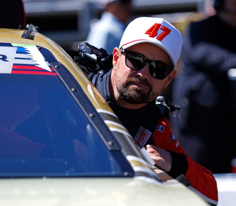 A man with sunglasses and a white cap with red '47' sitting in a race car, with a crowd blurred in the background.
