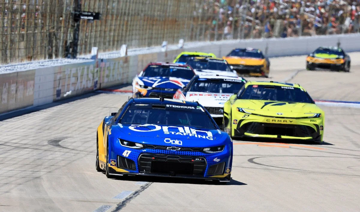 Race cars on a track, speeding and making a turn, with a barrier and audience in the background.