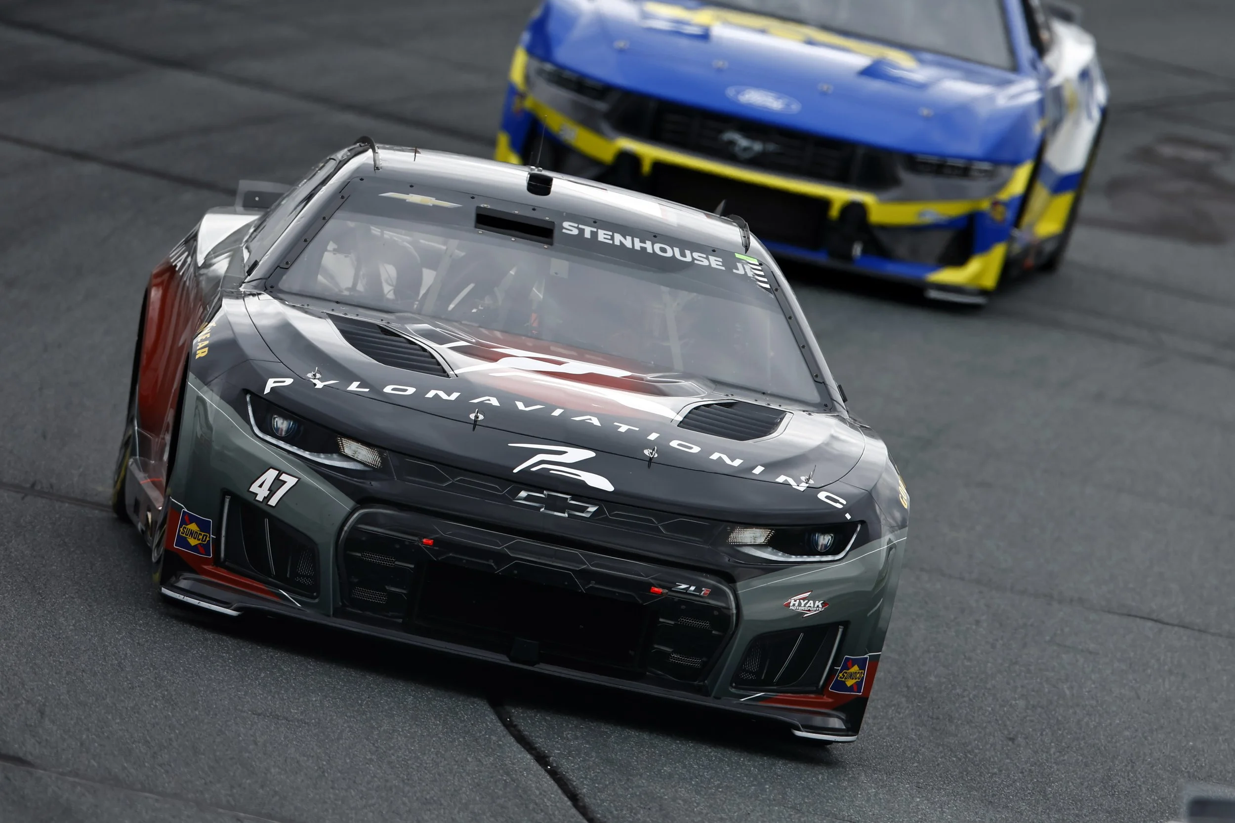 Close-up of a race car on a racetrack, with a second blue and yellow race car behind it. The cars are sleek, black and gray with sponsor logos and detailed aerodynamics, racing on a dark asphalt surface.