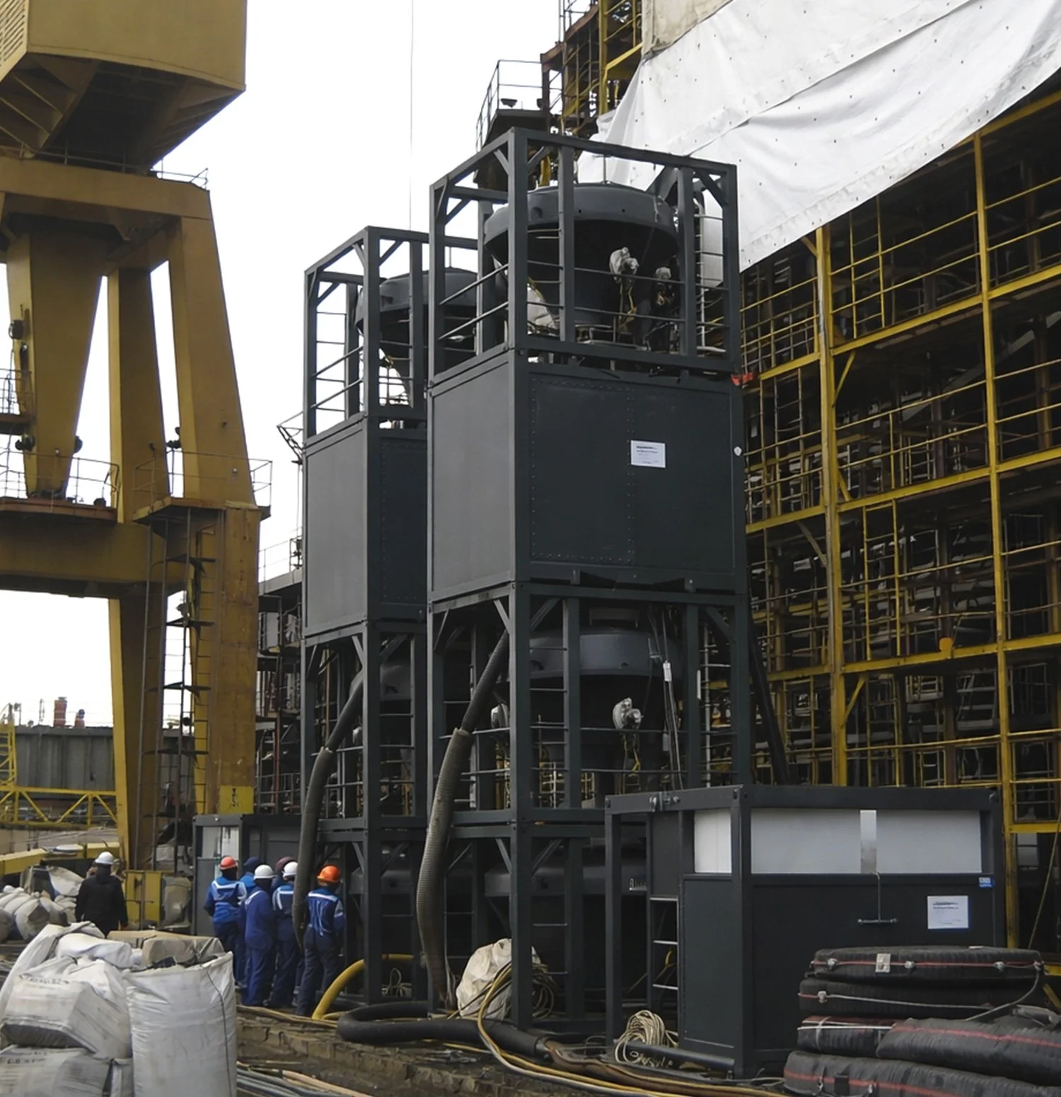 Construction site with workers and large industrial equipment, including a tall black structure with pipes and a control box, yellow cranes, and scaffolding in the background.