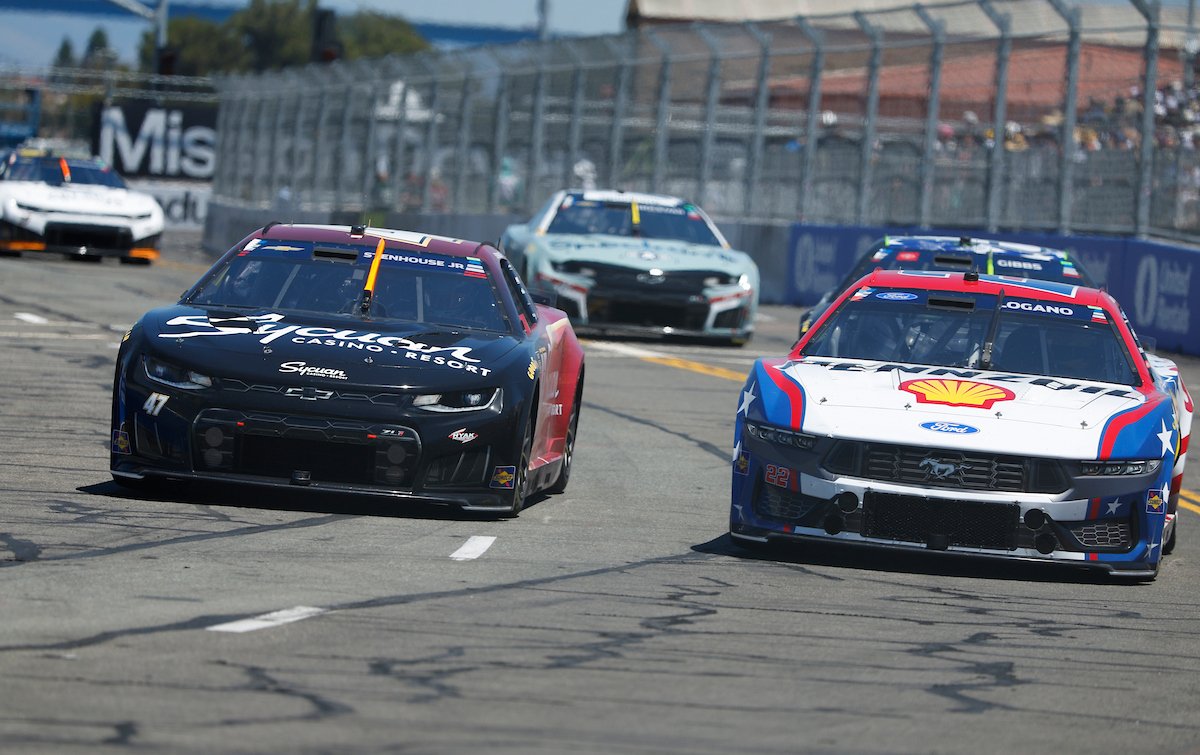 NASCAR race cars on a track, including a black car with red accents and sponsorship for Casino & Resort, and a white car with blue and red accents and a Shell logo, racing and kicking up dust.