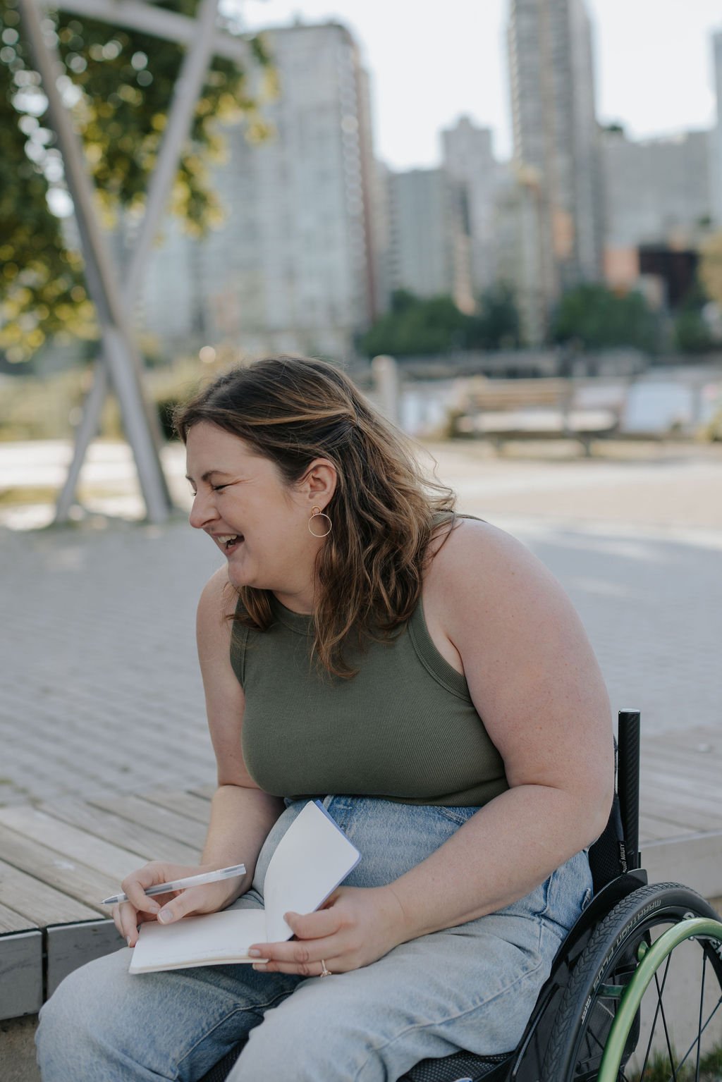 Ali is sitting in her wheelchair and laughing outdoors, holding a notebook and pen