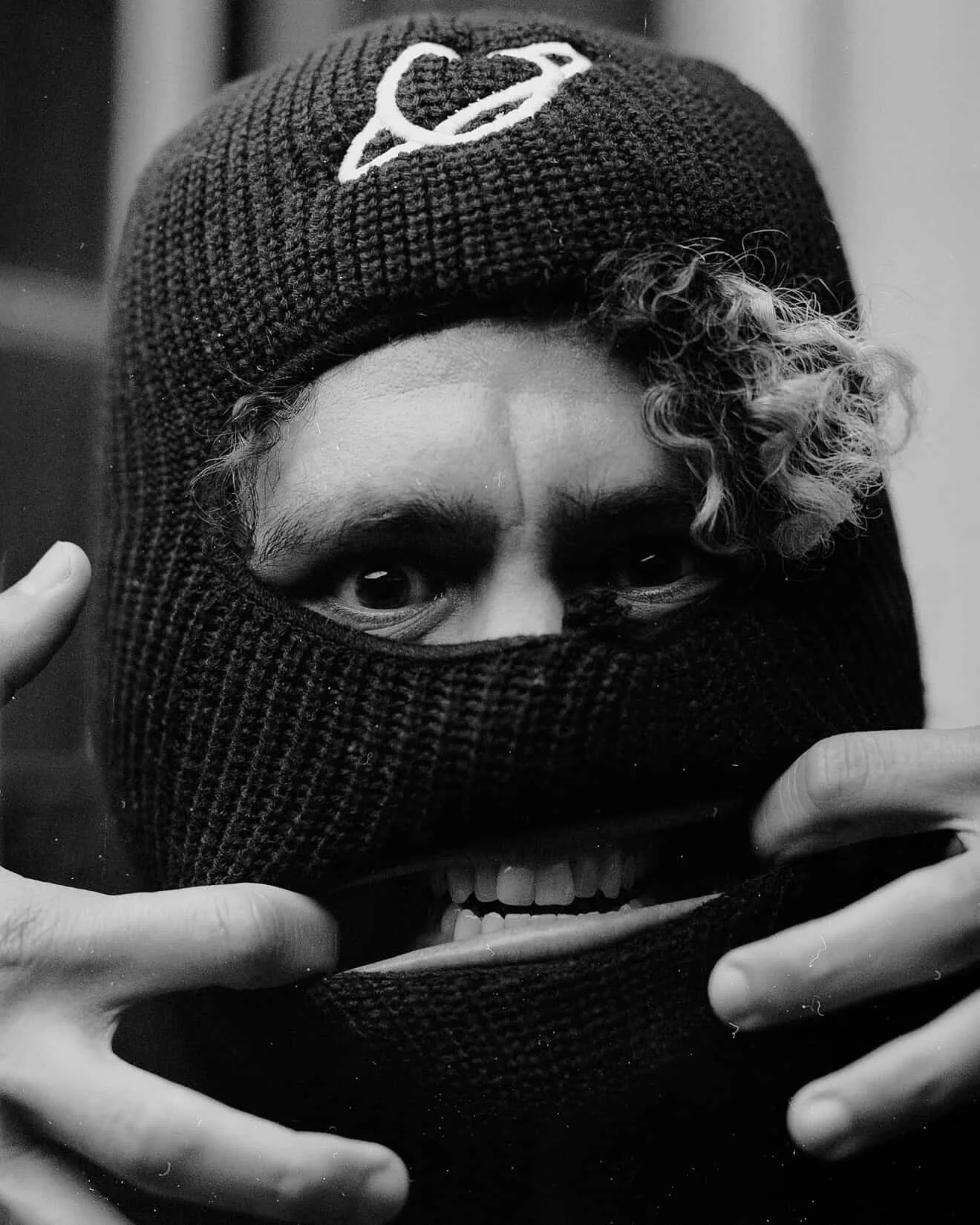 Person wearing a knit beanie and balaclava, smiling and making a gesture with their hands.