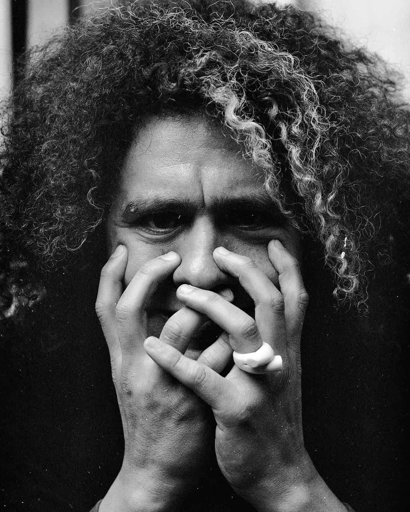 A close-up black and white photo of a woman with curly hair and dark eyes, covering part of her face with her hands, wearing a ring on her finger.