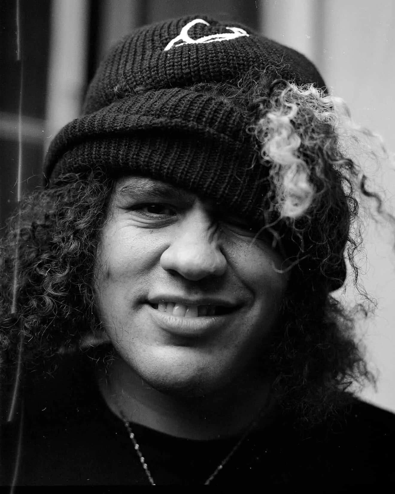 Black and white photo of a young person smiling, wearing a knit beanie and a curly hairstyle.