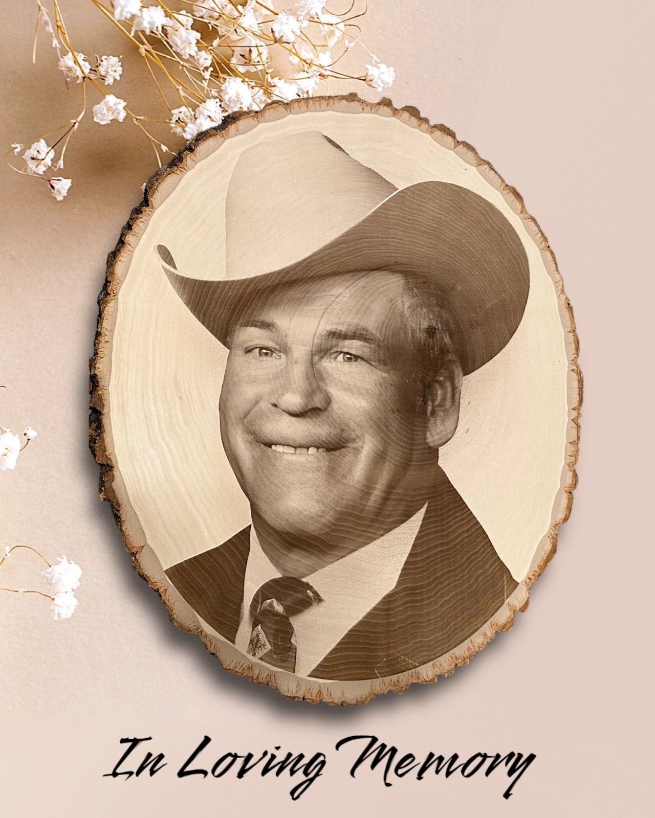 A laser-engraved wood slice features a portrait of an older man wearing a cowboy hat and a suit with a tie. The engraving is detailed and centered on a natural-edge wooden slab. The background is soft beige with delicate white baby’s breath flowers s
