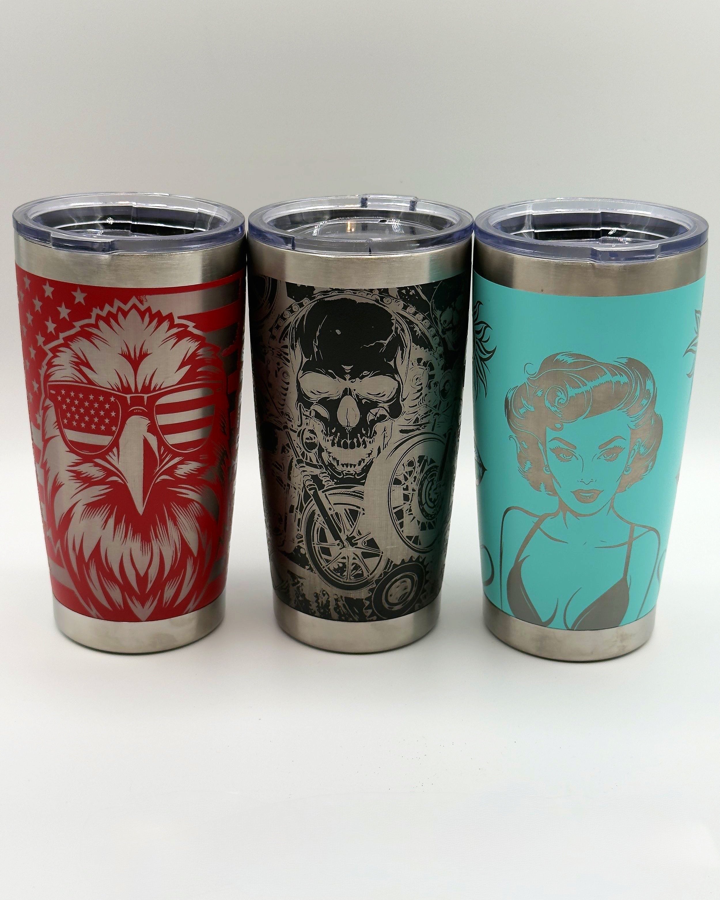Three custom laser-engraved tumblers are displayed side by side. The red tumbler on the left features an eagle wearing American flag sunglasses. The black tumbler in the center displays a menacing skull with motorcycle gears. The teal tumbler on the