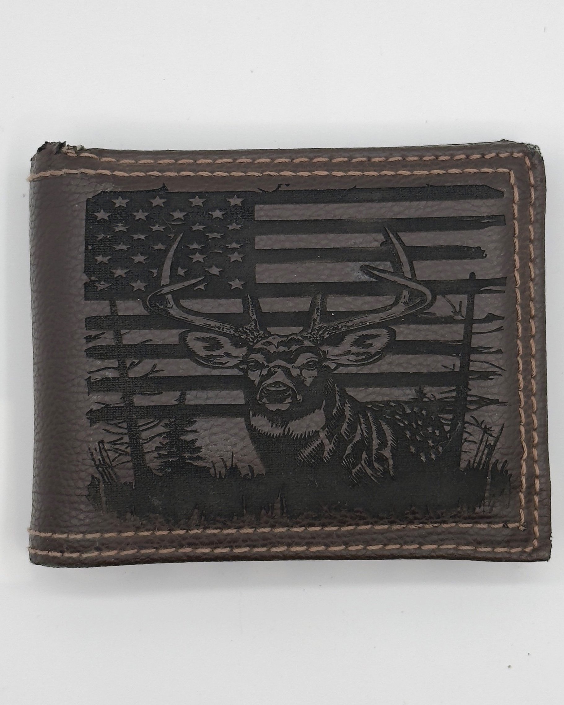 A brown leather wallet with tan stitching, laser engraved with a detailed design featuring a buck in the foreground and a distressed American flag in the background, flanked by silhouettes of trees. The engraving showcases fine contrast and texture,