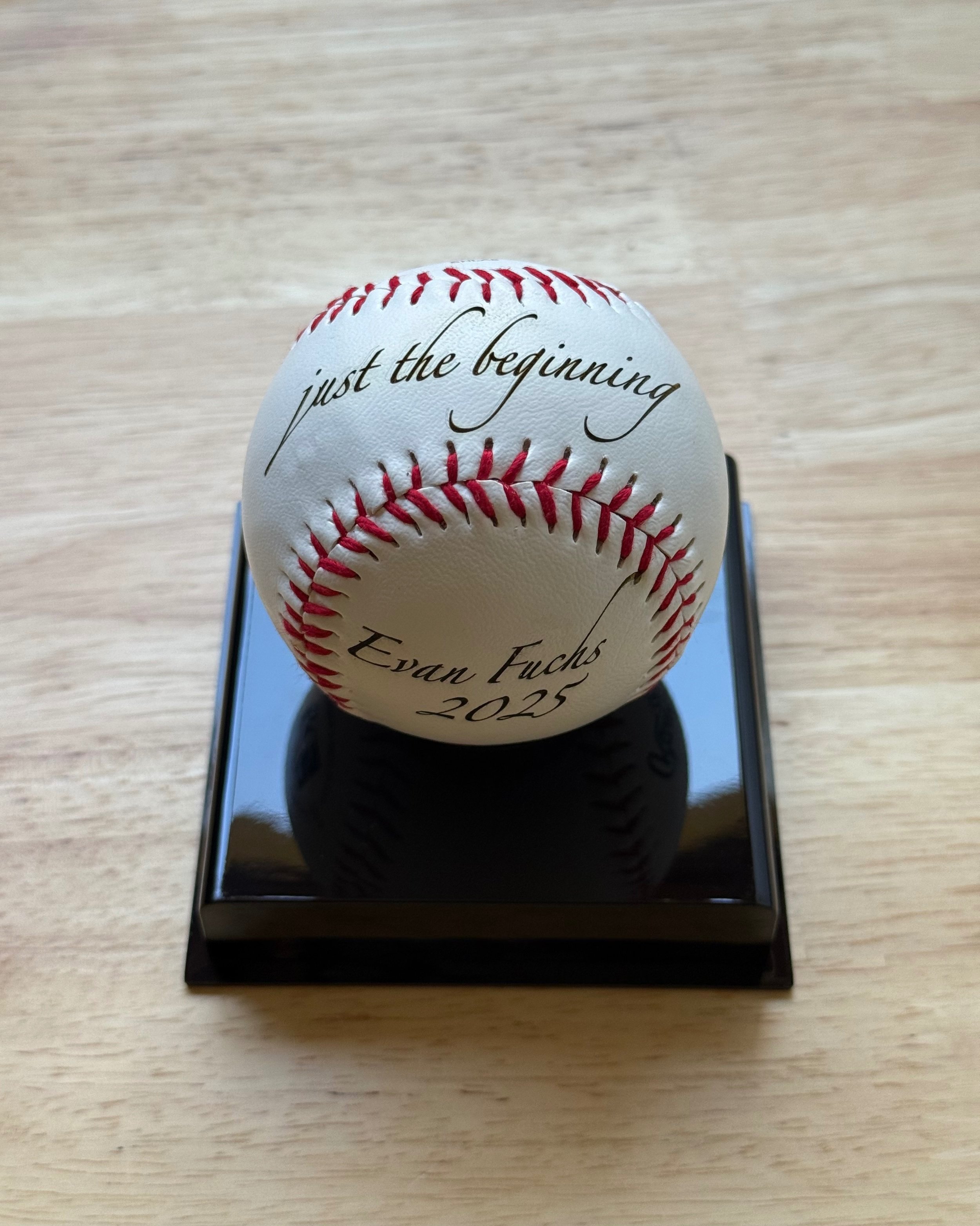 A white leather baseball with red stitching, engraved in elegant script with the words “just the beginning” on the top and “Evan Fuchs 2025” on the bottom. The ball is displayed on a glossy black stand, placed on a light wooden surface.