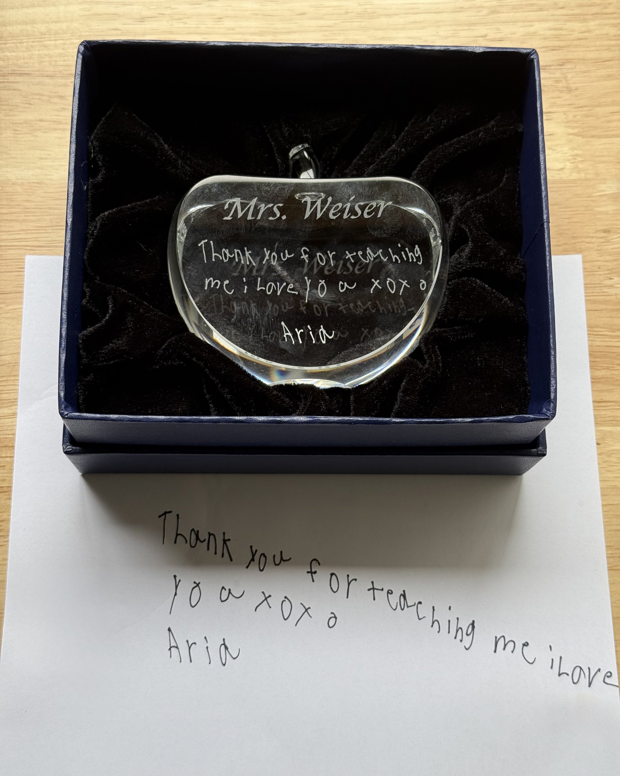 A crystal apple ornament rests in a velvet-lined gift box, engraved with a handwritten message that reads:
“Mrs. Weiser, Thank you for teaching me. I love you xoxo Aria.”
Beneath the box is the original note in a child’s handwriting, which was scanne