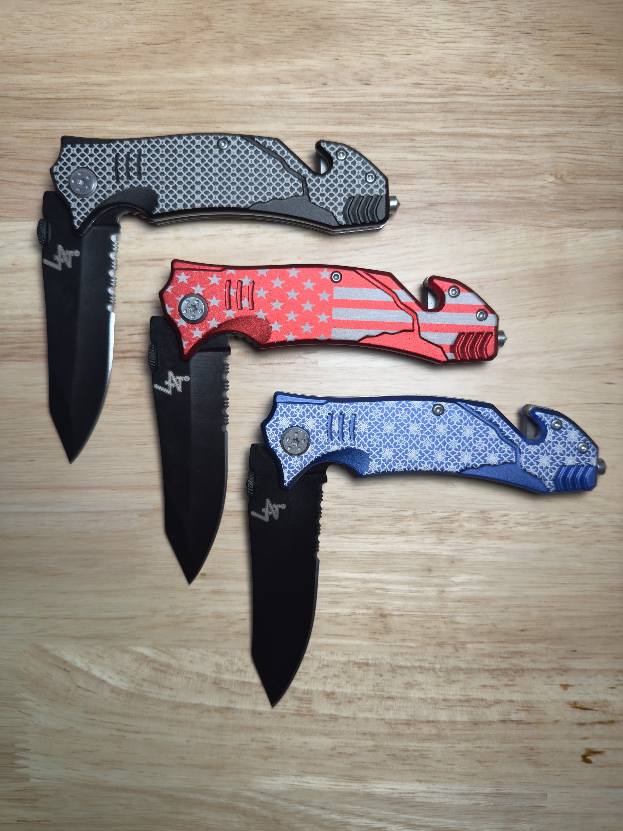 Three custom laser-engraved folding knives are arranged on a wooden surface. Each knife features a unique anodized aluminum handle: the top one is black with a hexagonal pattern, the middle is red with a stars and stripes American flag design, and th
