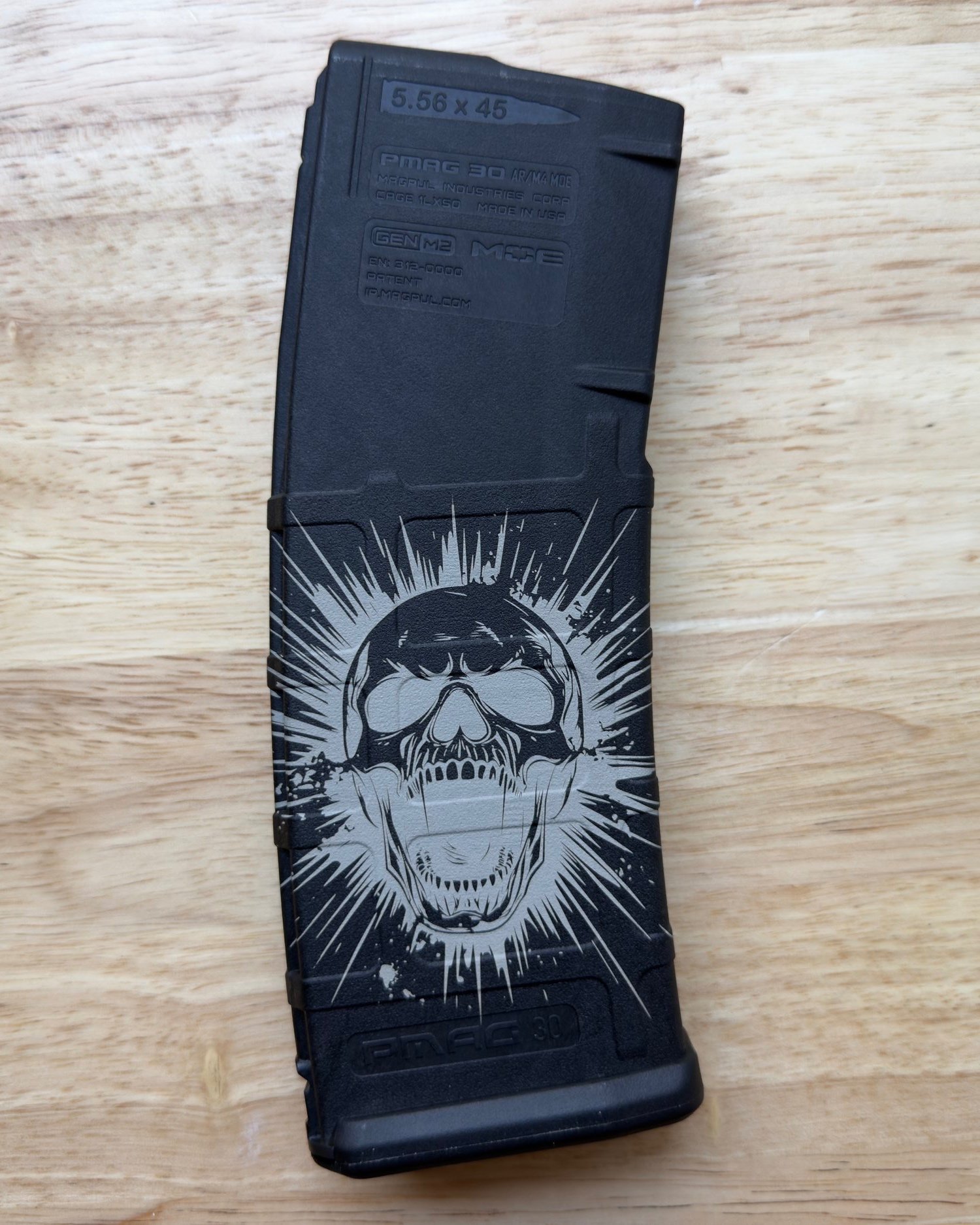A black Magpul PMAG 30-round magazine chambered for 5.56×45mm is shown with a custom laser engraving on the lower half. The design features a stylized skull wearing sunglasses, surrounded by an explosive burst pattern that radiates outward, giving th