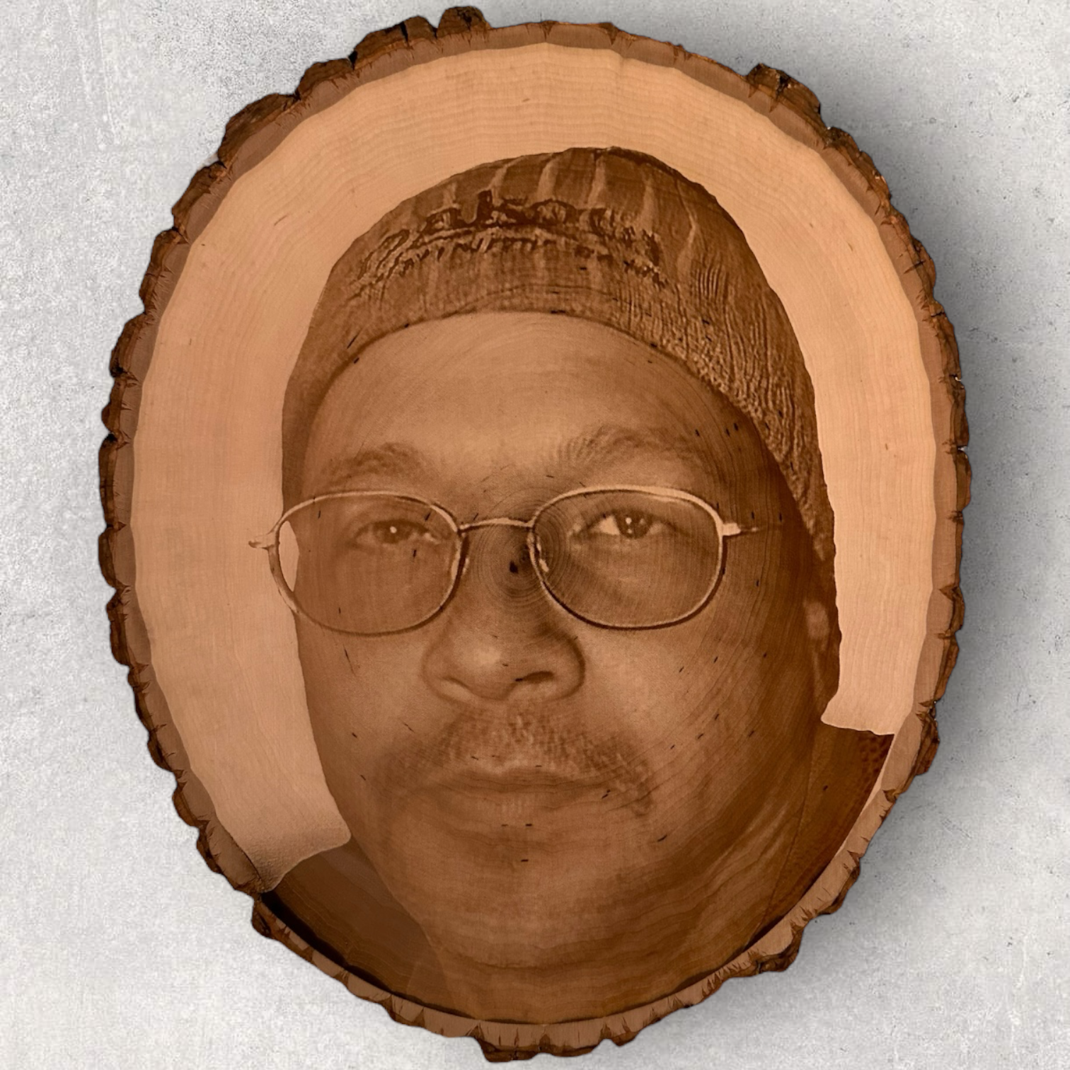 Custom laser engraved portrait on oval live edge basswood slab featuring a man wearing glasses and a beanie, etched with natural bark border on light wood background - Lazeric