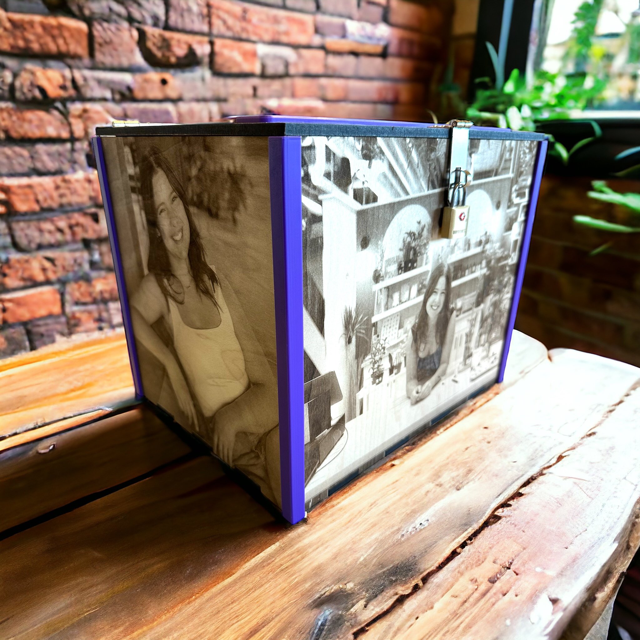 A custom laser-engraved event card box featuring four black-and-white engraved photos mounted on wood panels, framed with purple 3D-printed corner accents. The box is shown on a rustic wooden table with a brick wall and greenery in the background. A