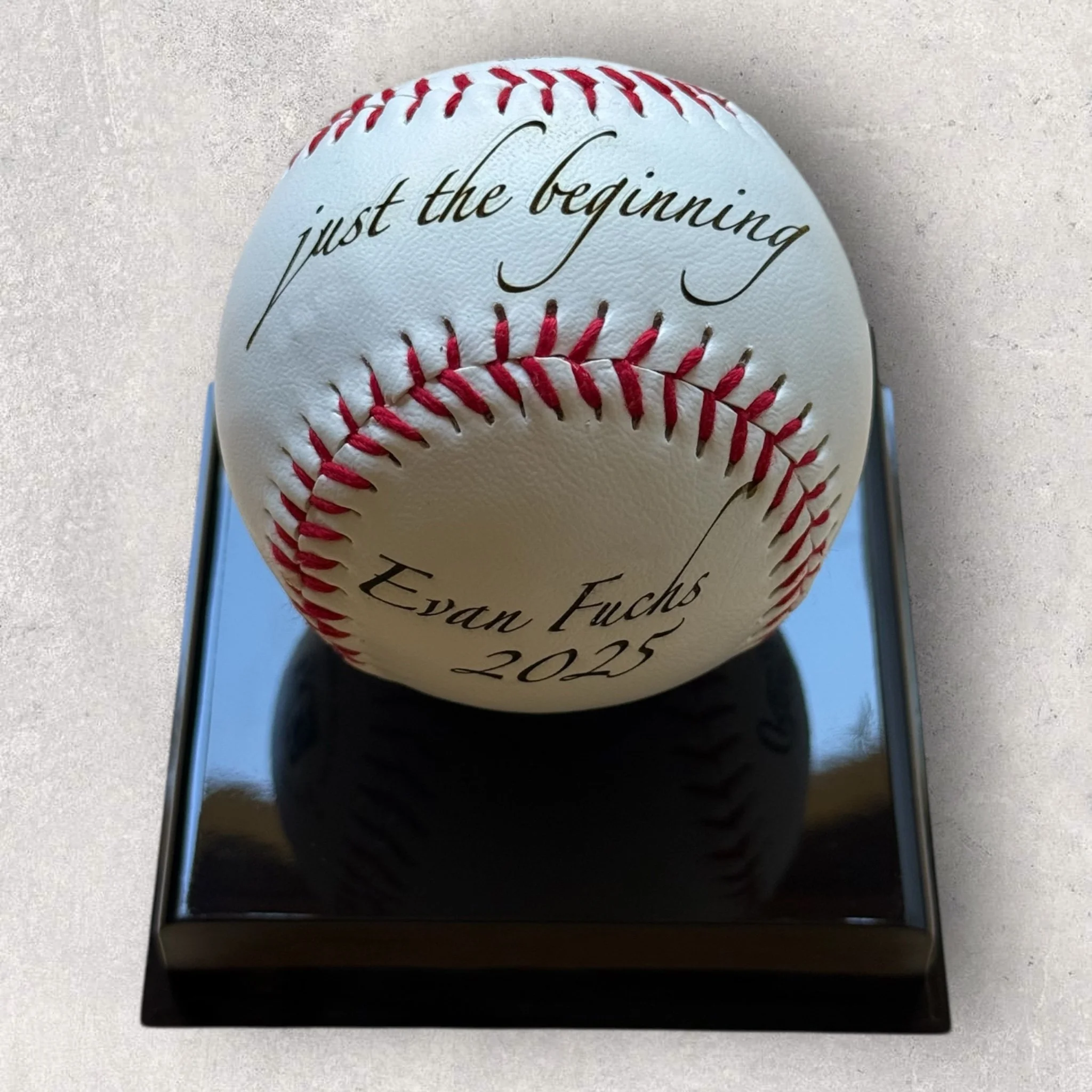 Laser-engraved baseball displayed on a black stand, featuring the phrase ‘just the beginning,’ a personalized name, and year, created as a custom sports keepsake by Lazeric.