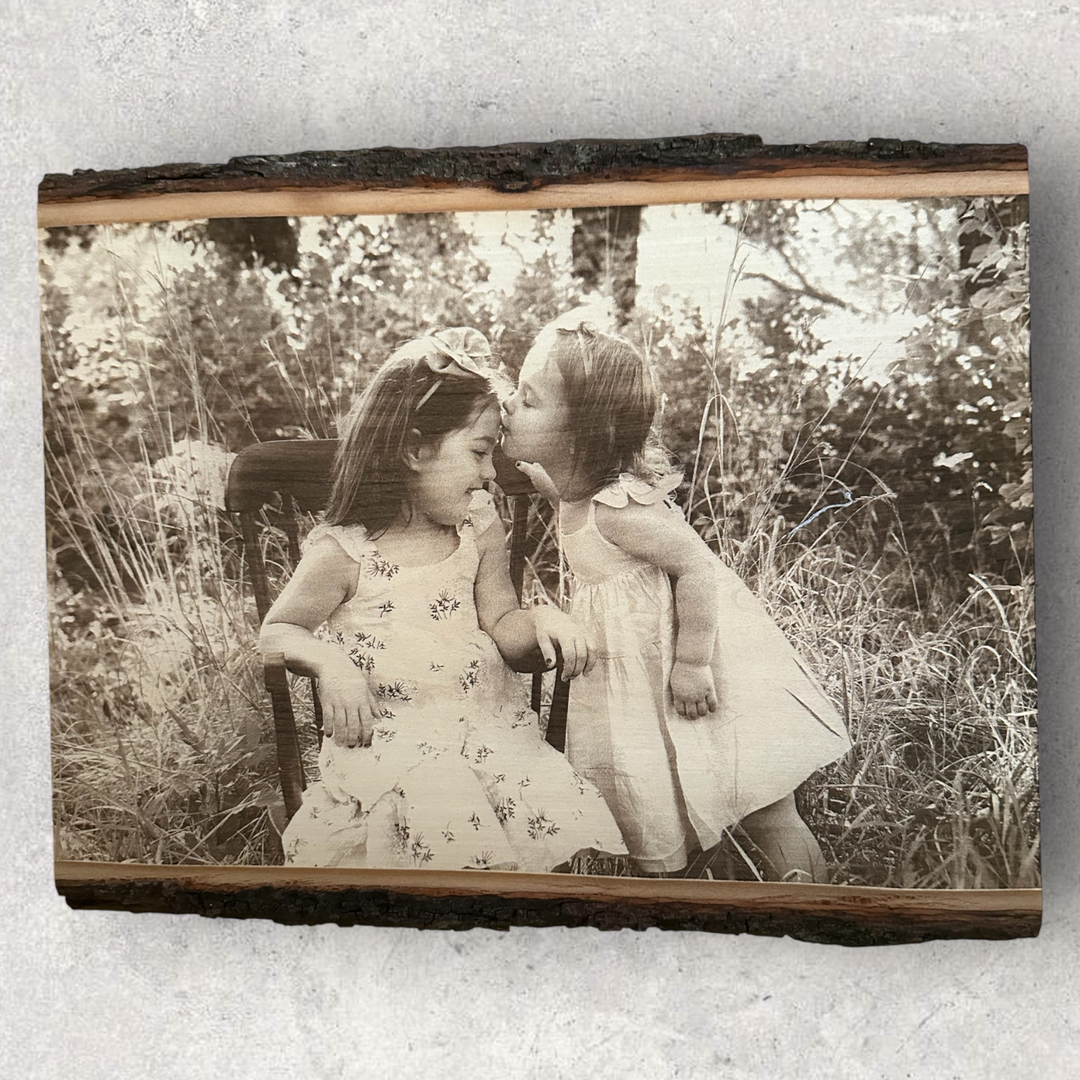 Laser engraved photo of two young sisters sharing a kiss, etched on rectangular live edge basswood with visible bark and natural wood grain, displayed on a light concrete surface