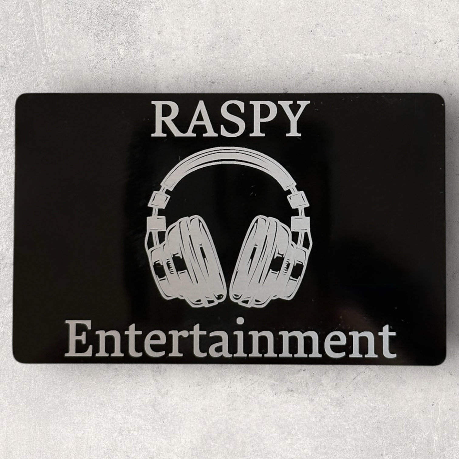 Laser engraved metal business card for Raspy Entertainment with bold text and detailed headphone logo - Lazeric