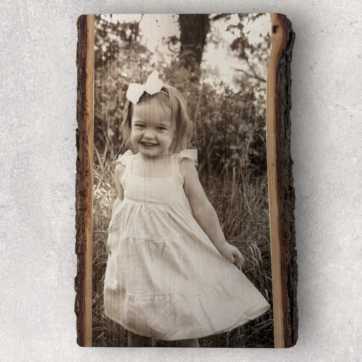 Laser engraved photo of a smiling toddler in a white dress standing outdoors, on bark-edged hardwood