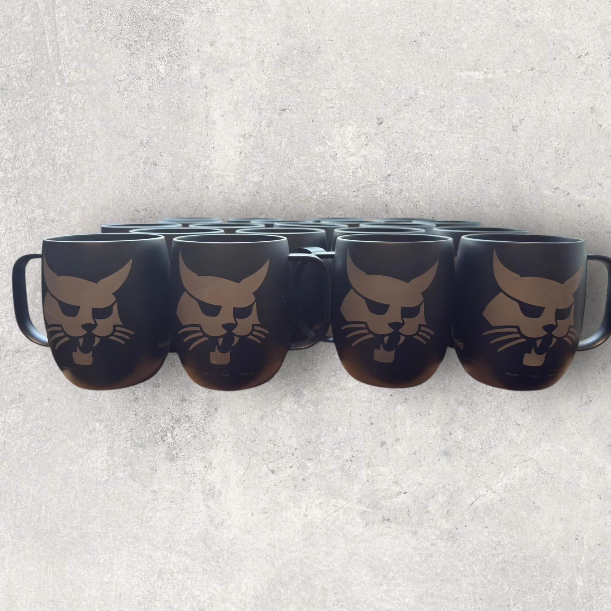 Custom laser-engraved self-heating coffee mugs with the Bobcat logo, shown as a bulk corporate order of black smart mugs engraved for employee gifts and branded promotional use.