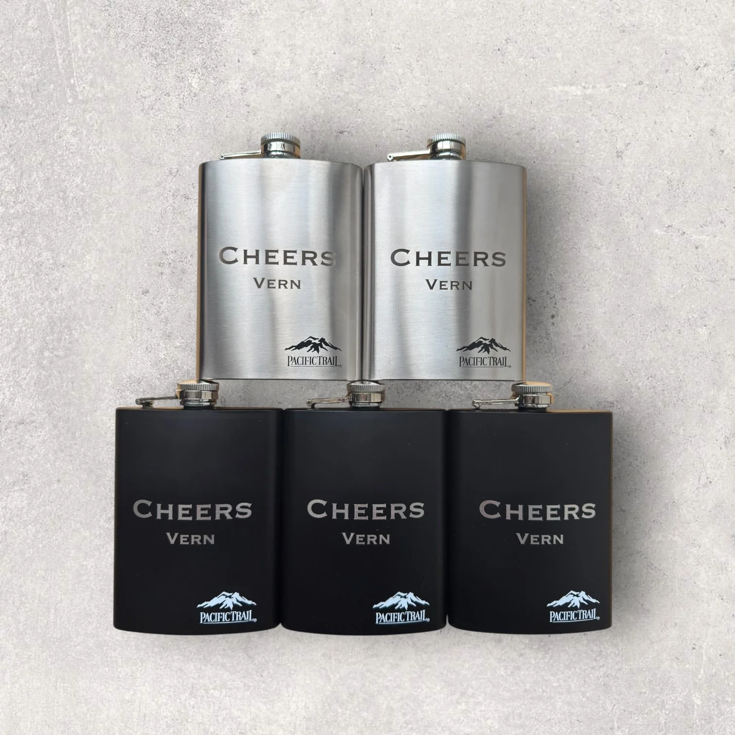 Set of laser-engraved stainless steel flasks with custom names and logo, featuring both brushed silver and matte black finishes for personalized gift engraving.