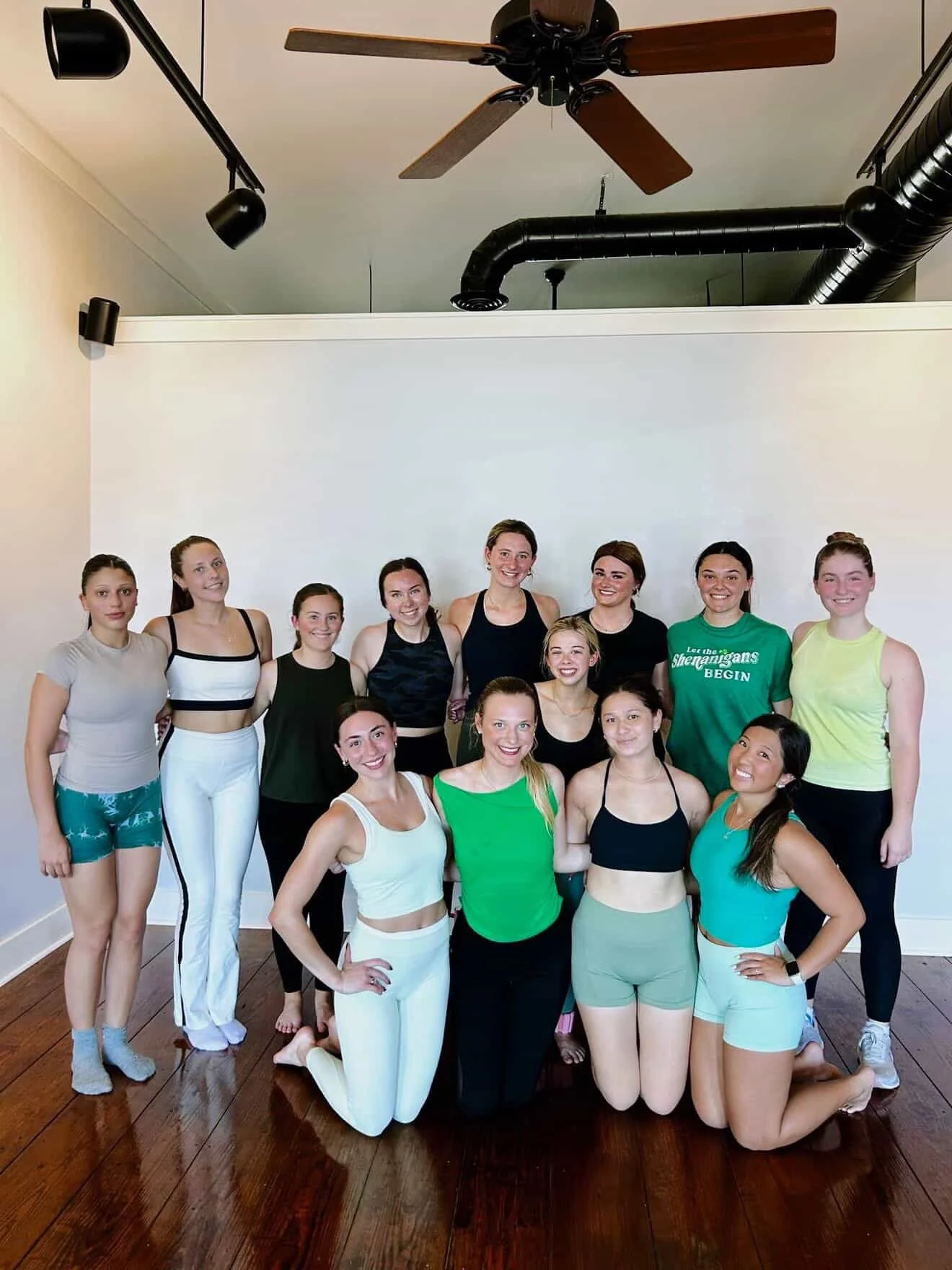 A group of 14 women dressed in athletic wear, posing together indoors with a white wall background, wooden floor, ceiling fan, and black industrial air ducts.