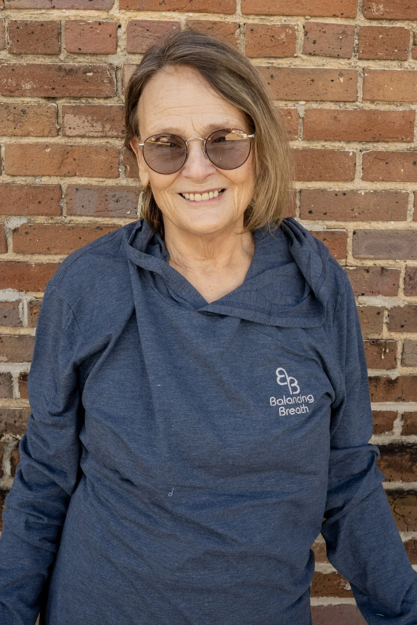 A smiling woman wearing glasses and earrings, standing against a brick wall.