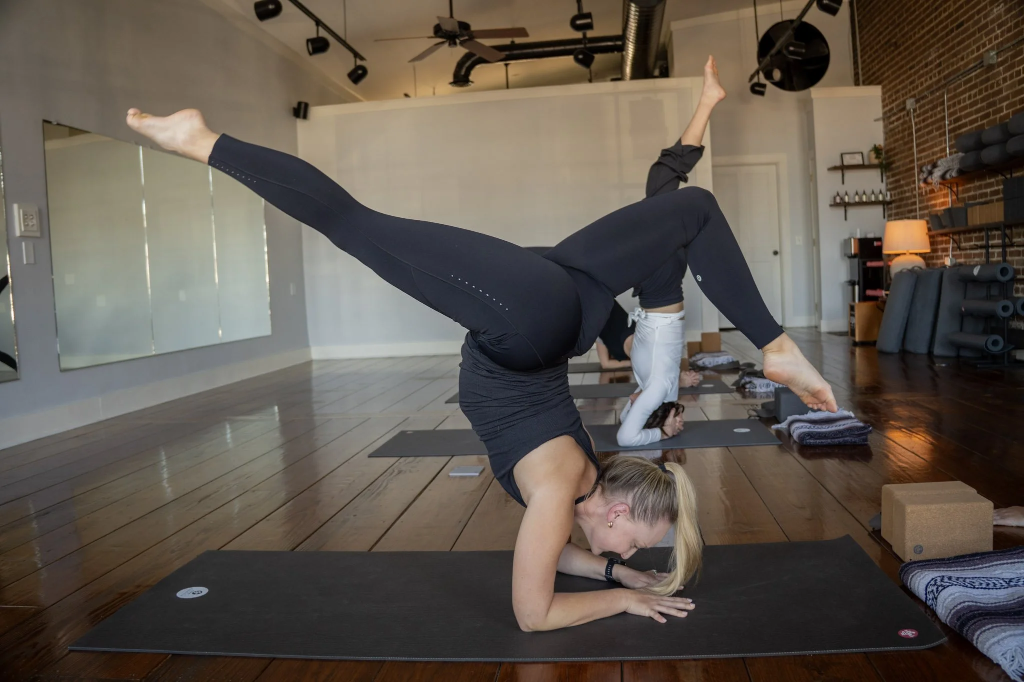 A woman practicing yoga in a studio, balancing on her forearms with one leg lifted straight back and the other bent, surrounded by yoga mats and equipment.