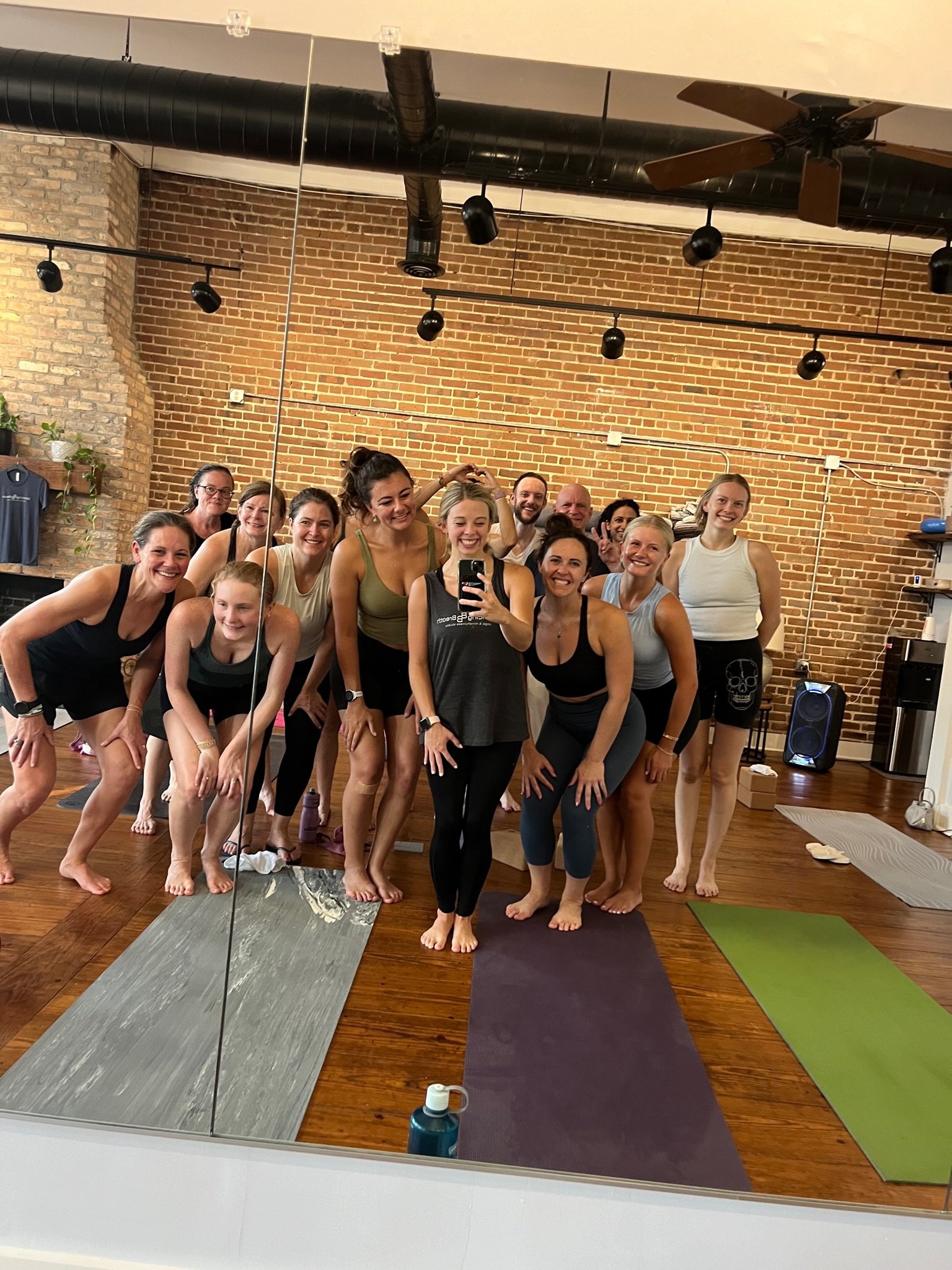 Group of people in workout clothes taking a selfie in a fitness studio with a brick wall background and yoga mats.