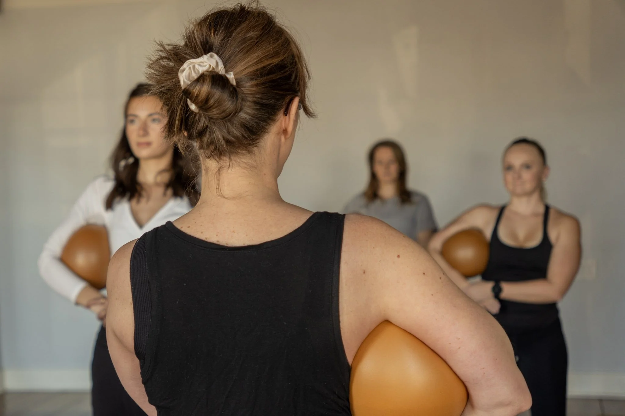Two women participating in a fitness class, holding small exercise balls against their chests, in a studio with wooden floors and a mirror.