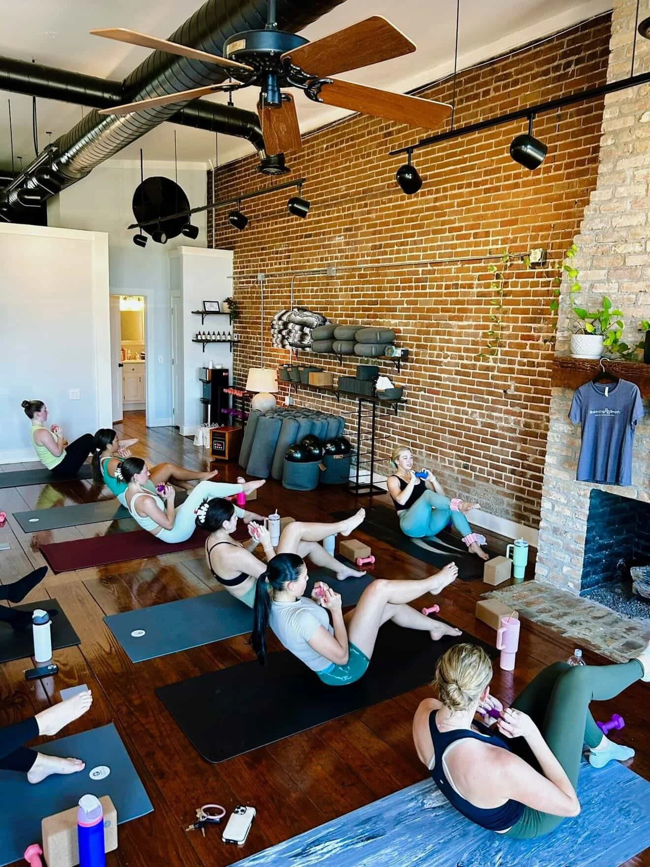 Women participating in a yoga or fitness class in a studio with brick walls, hardwood floors, and a ceiling fan.