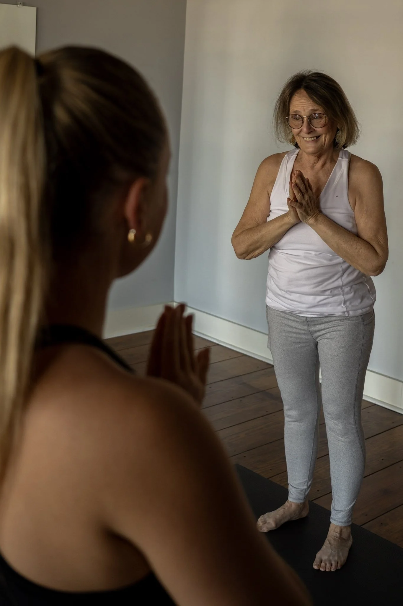 An elderly woman with glasses and gray hair smiling with hands pressed together in a gesture of greeting or gratitude, facing a younger woman practicing yoga or meditation in a room with wooden floors.